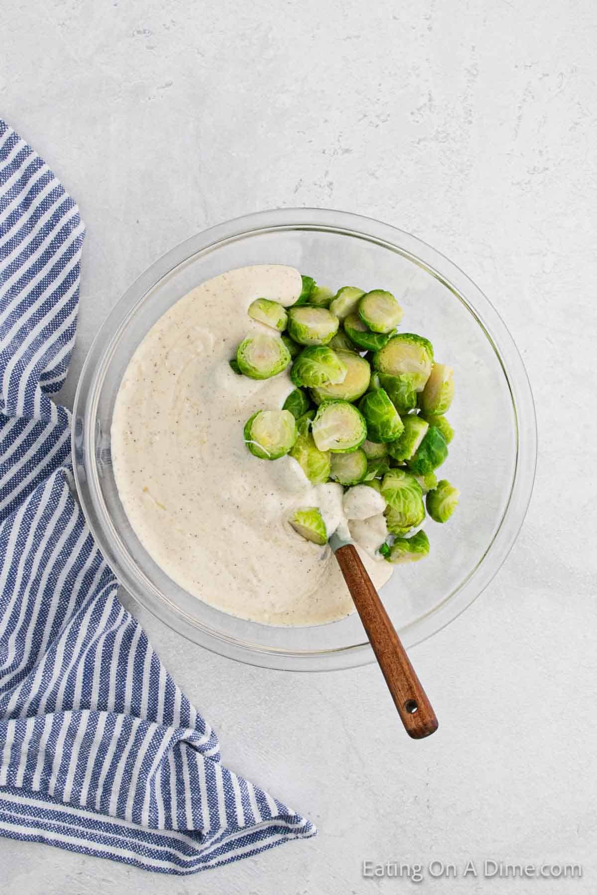 A glass bowl of creamy dressing with halved Brussels sprouts au gratin on top. A wooden spoon is resting in the bowl. A blue and white striped cloth lies beside it, evoking a rustic charm to this elegant recipe. The scene is set against a light gray stone surface.