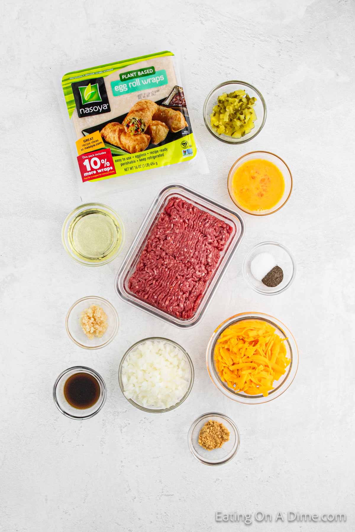 A package of plant-based egg roll wraps sits alongside bowls of cheeseburger-inspired ingredients: ground meat, shredded cheese, diced onions, garlic, mustard, Worcestershire sauce, beaten egg, and pickles. The backdrop is a light gray surface.