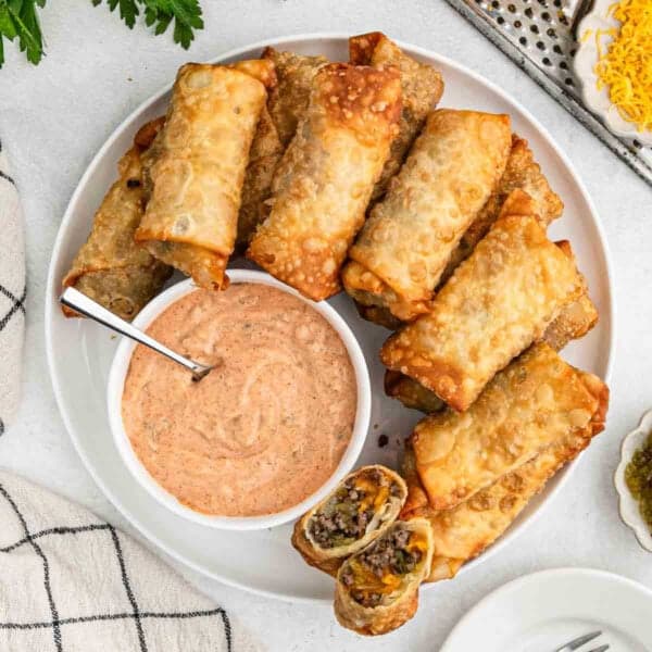 A white plate filled with cheeseburger-inspired egg rolls, some split open to reveal a beef and cheese filling. A small bowl of creamy orange dip with a spoon is also on the plate. Nearby are a green towel and a plate with cheese and pickles.