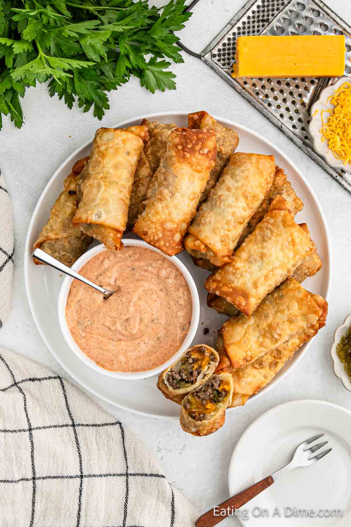 A plate of golden-brown egg rolls with a cheeseburger twist, accompanied by a zesty dipping sauce and a spoon in a bowl. The dish is garnished with fresh parsley, next to a checkered kitchen towel and a block of cheese on a grater, surrounded by shredded cheese and pickles.