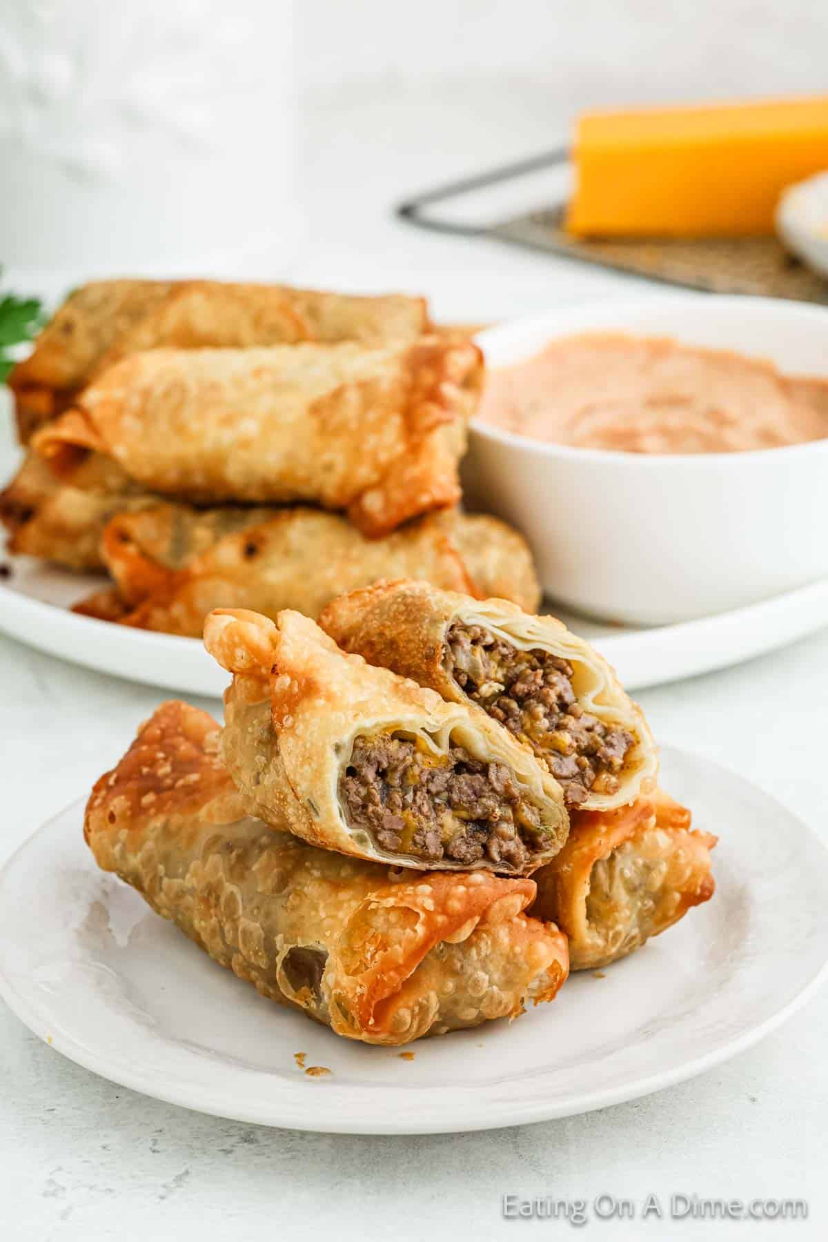 A plate with two crispy cheeseburger egg rolls, one cut open to reveal the savory beef filling. In the background, more of these delicious snacks are stacked on a plate beside a bowl of dipping sauce.