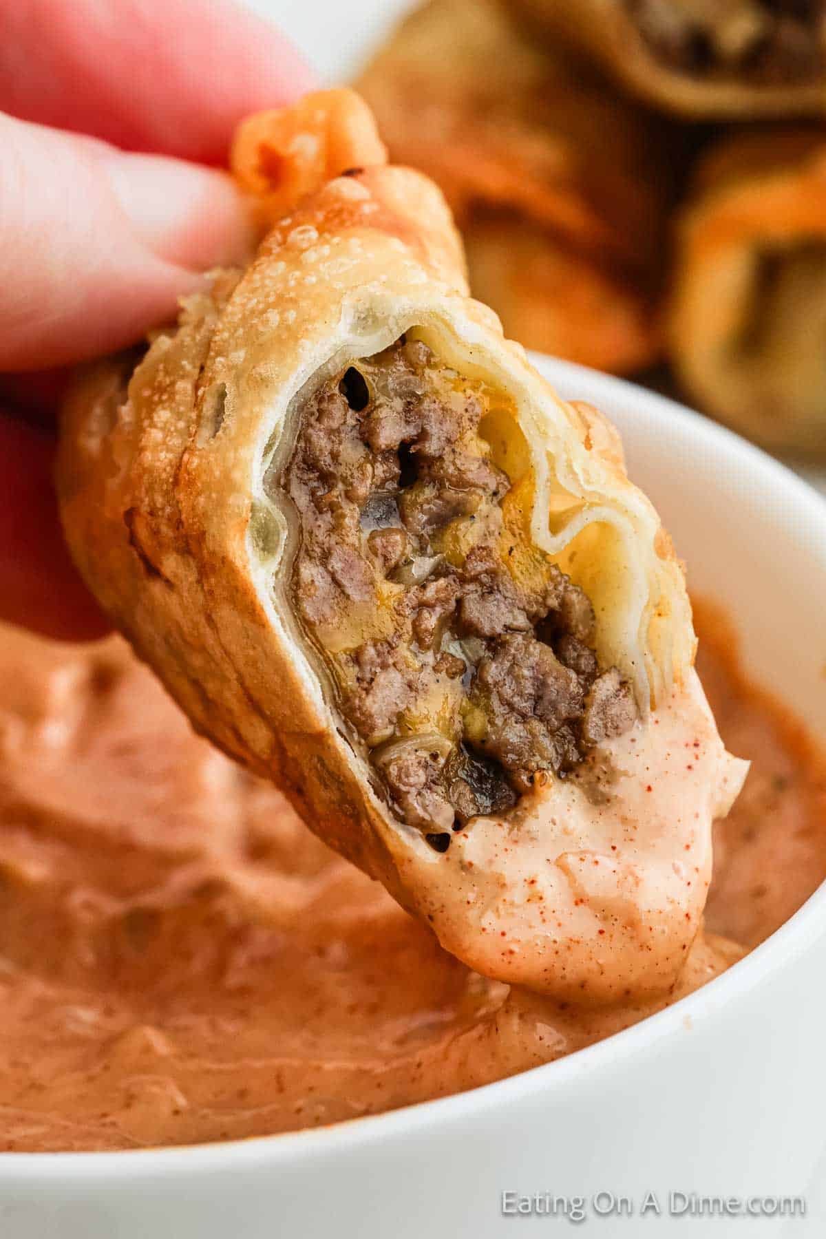 A close-up of a hand holding a pastry filled with meat and onions, reminiscent of a cheeseburger. The pastry is partially dipped in a creamy, spiced sauce, adding an extra layer of flavor. Another piece of this delectable treat is blurred in the background.