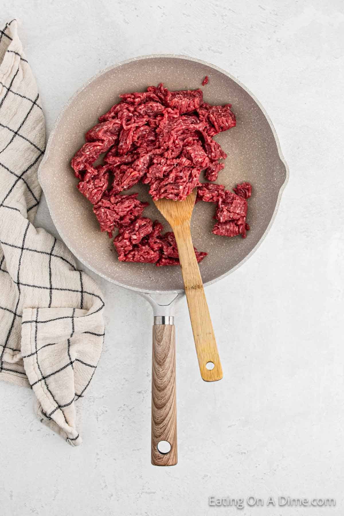 Top view of raw ground beef in a skillet with a wooden spatula, ready to transform into delightful cheeseburger patties, placed on a light surface next to a folded checkered kitchen towel.