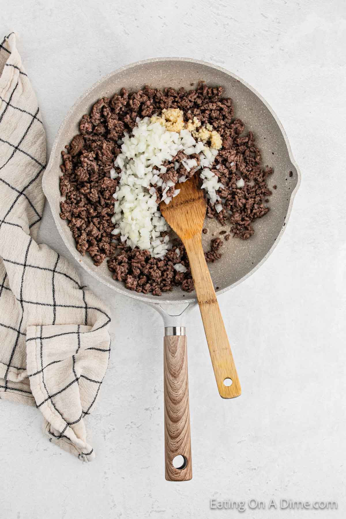 A frying pan on a gray surface contains cooked ground beef mixed with chopped onions and minced garlic, reminiscent of a hearty cheeseburger. A wooden spatula rests in the pan. To the left, a checkered kitchen towel is placed on the counter, completing the classic kitchen scene.