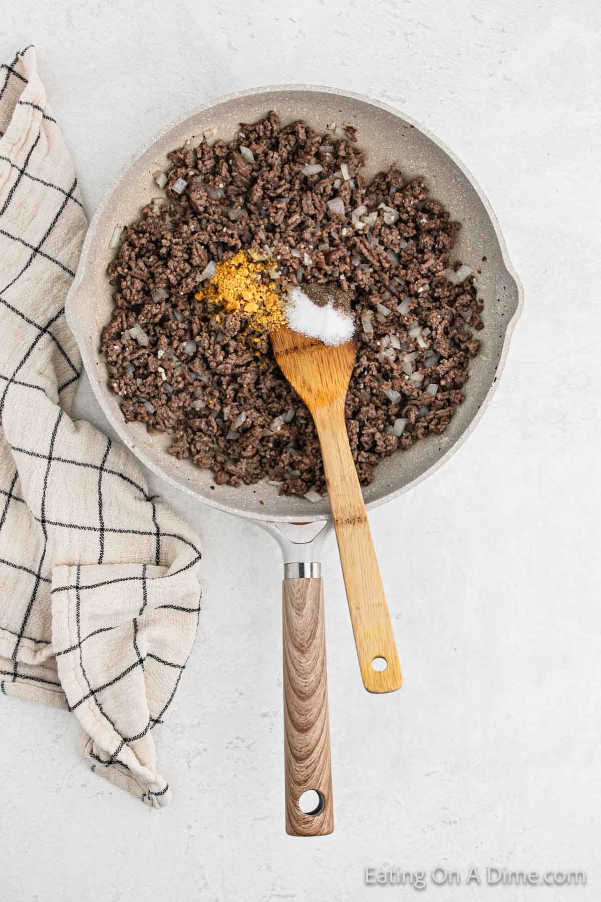 A frying pan filled with cooked ground beef and chopped onions, topped with a pile of seasoning and salt, sets the stage for delicious cheeseburger egg rolls. A wooden spoon rests in the pan, while a black and white checkered cloth lies beside it on a light gray surface.