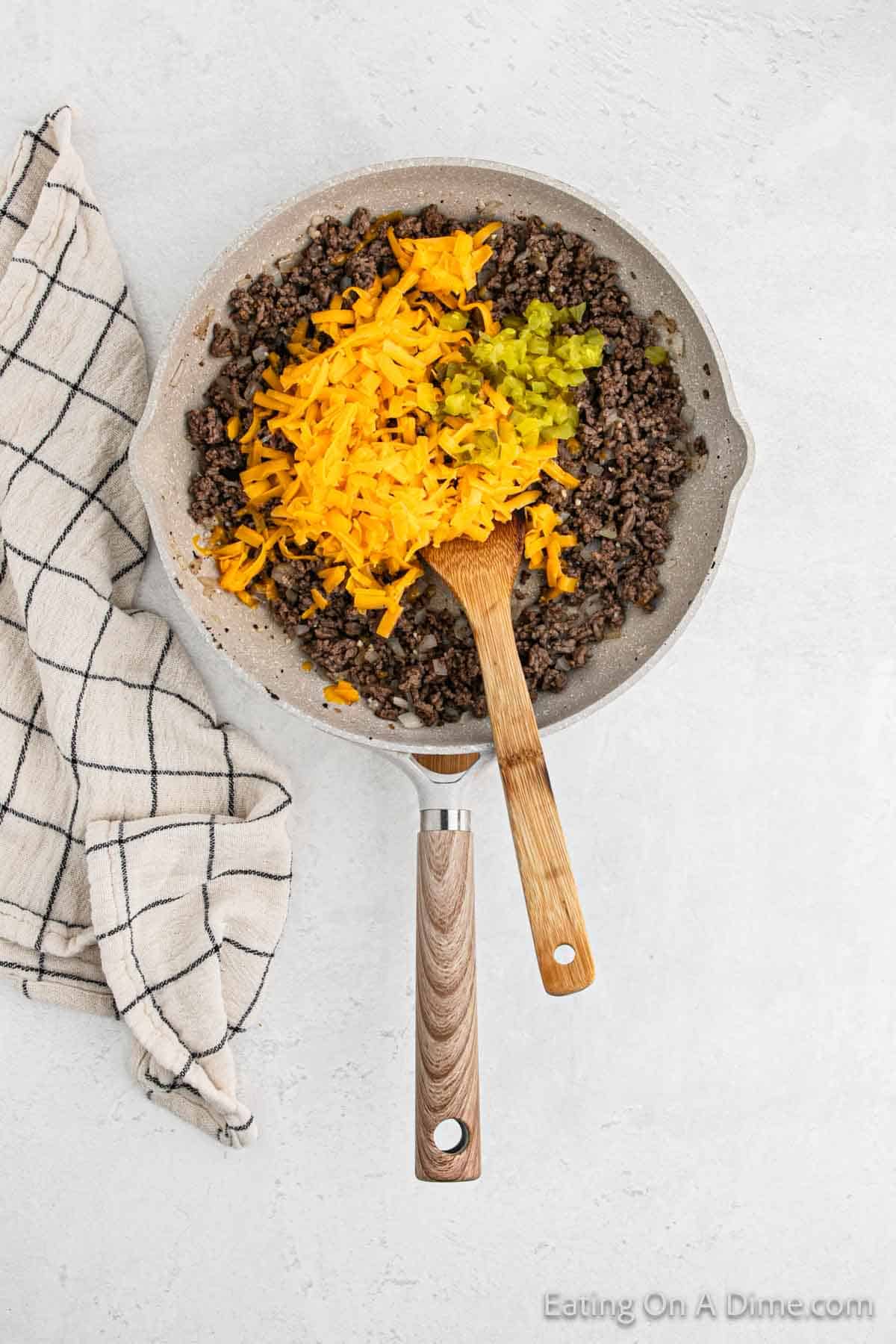 A skillet filled with savory ground meat, reminiscent of a deconstructed cheeseburger, is topped with melted cheese and chopped green peppers. A wooden spatula rests in the skillet, while a black and white checkered cloth lies beside it on the light countertop.