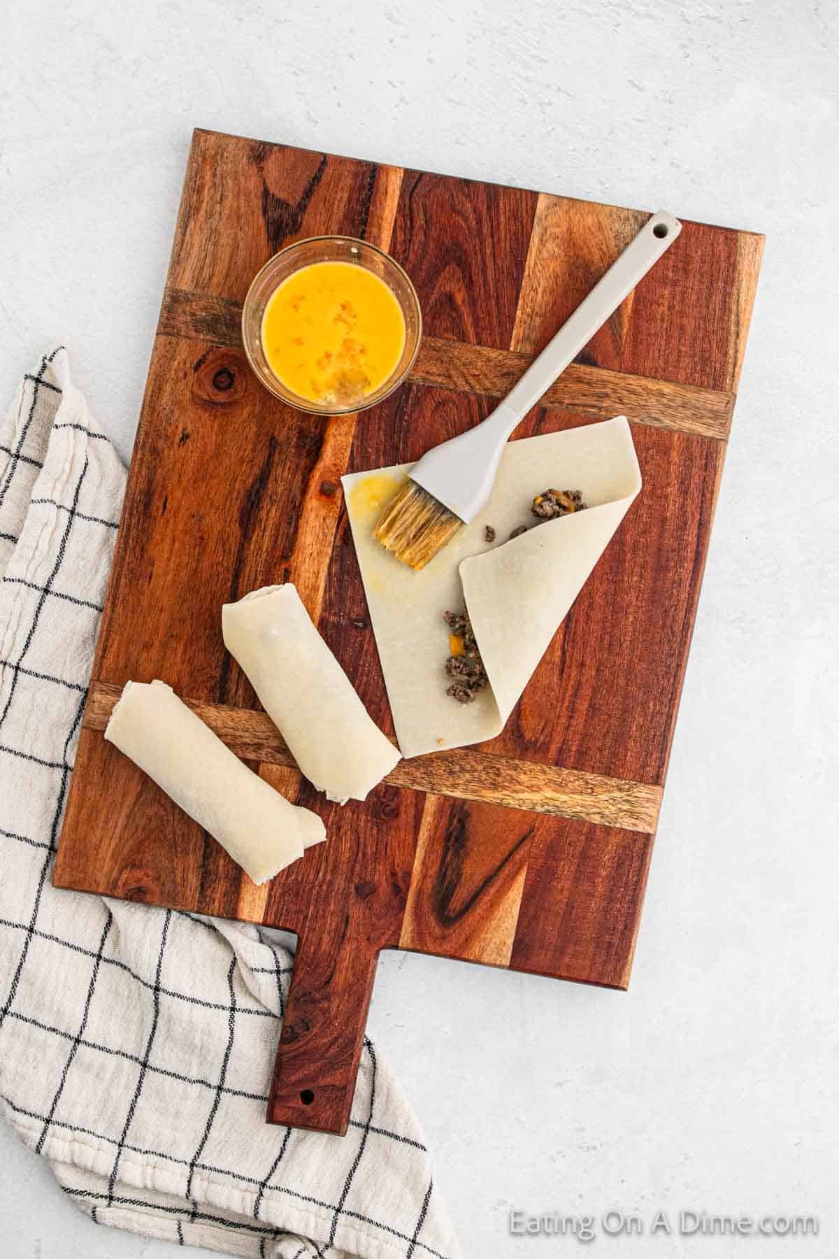 A wooden cutting board showcases three partially rolled egg rolls containing a savory filling. A small bowl of beaten egg and a brush are ready to seal the edges. A checkered cloth adds a rustic touch beside them on the light gray surface, hinting at cheeseburger-inspired flavors within.