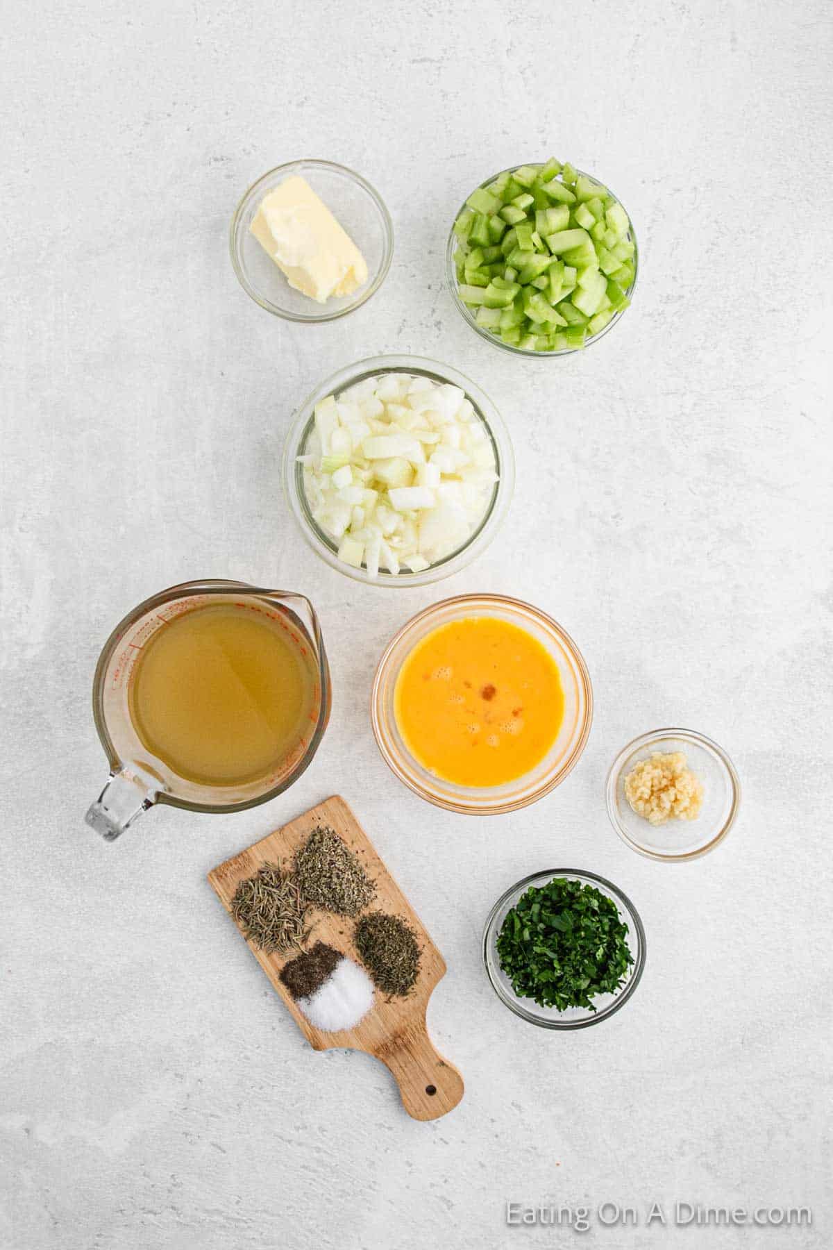 A delightful array of ingredients for cornbread stuffing awaits: diced green peppers, chopped onions, and minced garlic beside butter. Eggs and vegetable broth add richness, while thyme, salt, and pepper rest on a small wooden board—all beautifully arranged on a pristine white surface.