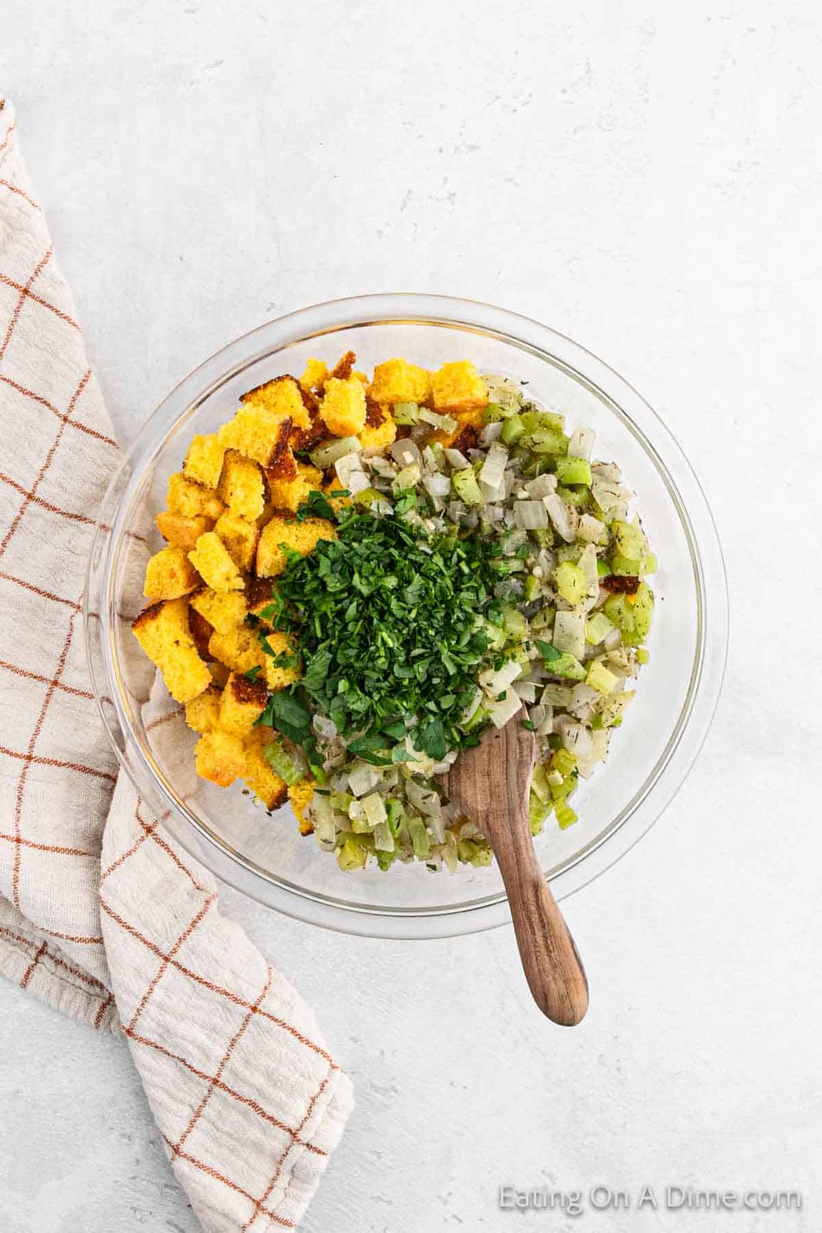 A glass bowl brimming with cornbread stuffing, featuring diced vegetables and chopped herbs, rests on a light surface. A wooden spoon peeks out, ready for serving, with a beige checkered cloth gracefully draped beside it.