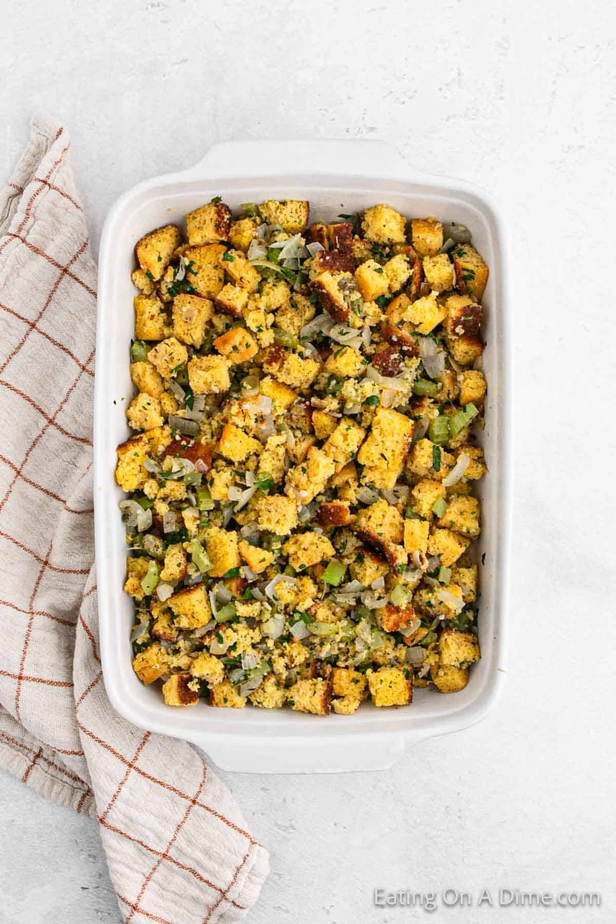 A baking dish filled with fluffy cornbread stuffing featuring golden-brown croutons, chopped celery, onions, and herbs. A beige and red checkered cloth is placed beside the dish on a light gray surface.