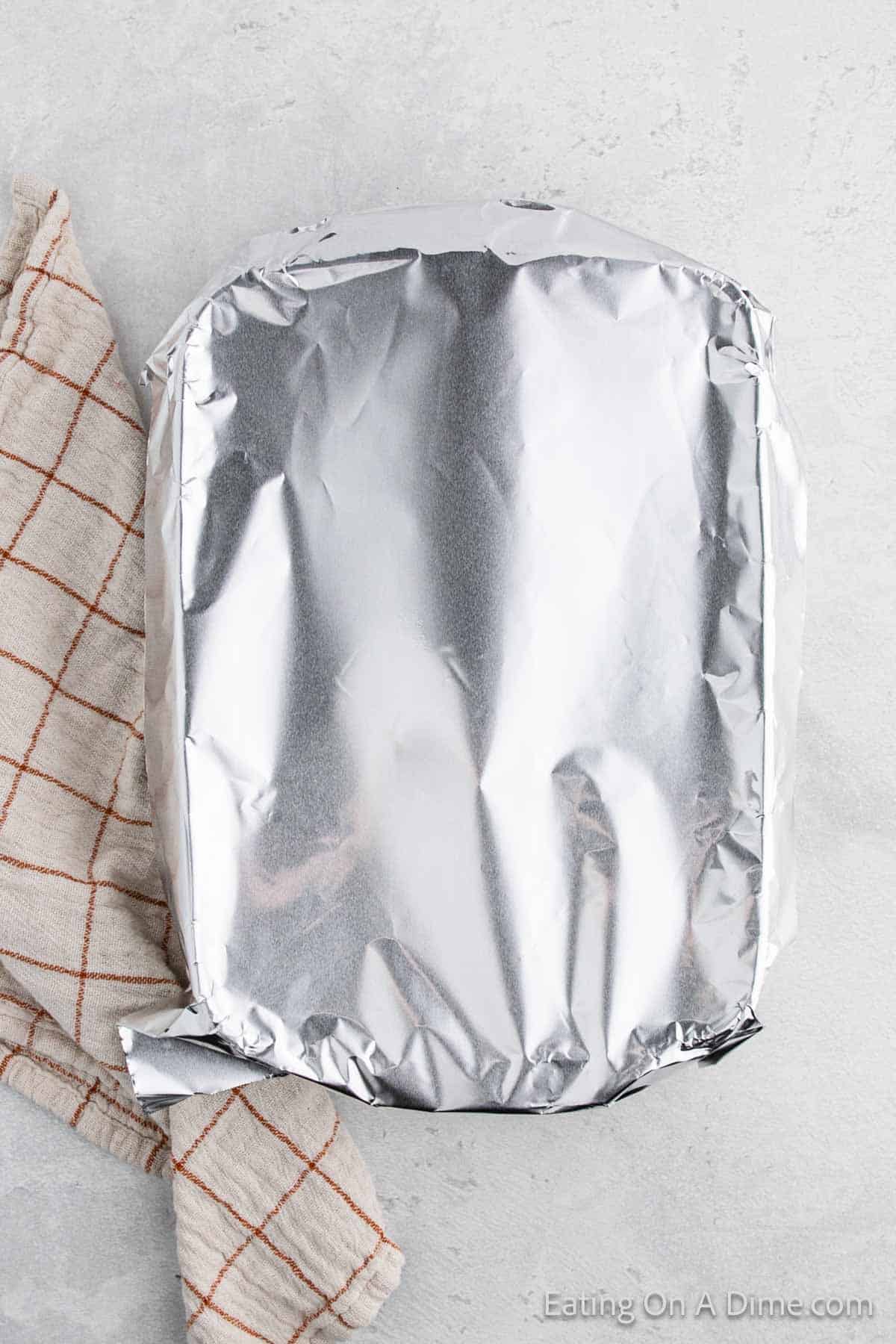 A dish filled with savory stuffing, covered in shiny aluminum foil, rests on a light surface next to a folded, beige cloth with red checkered lines.