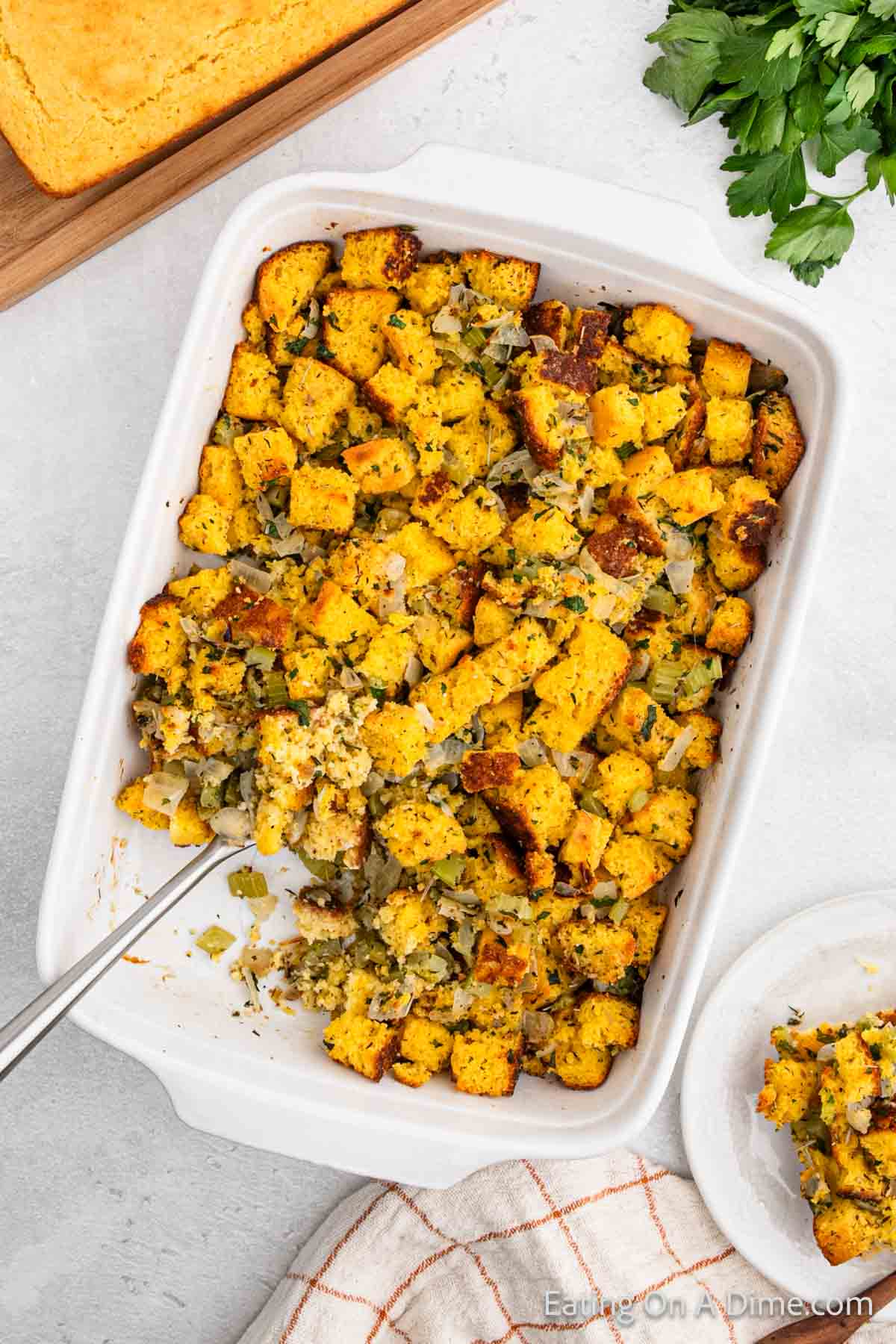 A baking dish filled with golden brown cornbread stuffing, topped with herbs and dotted with chopped onions. A serving spoon has scooped a portion onto a small white plate beside it. Fresh parsley and a tray of warm cornbread are in the background, adding to the inviting scene.