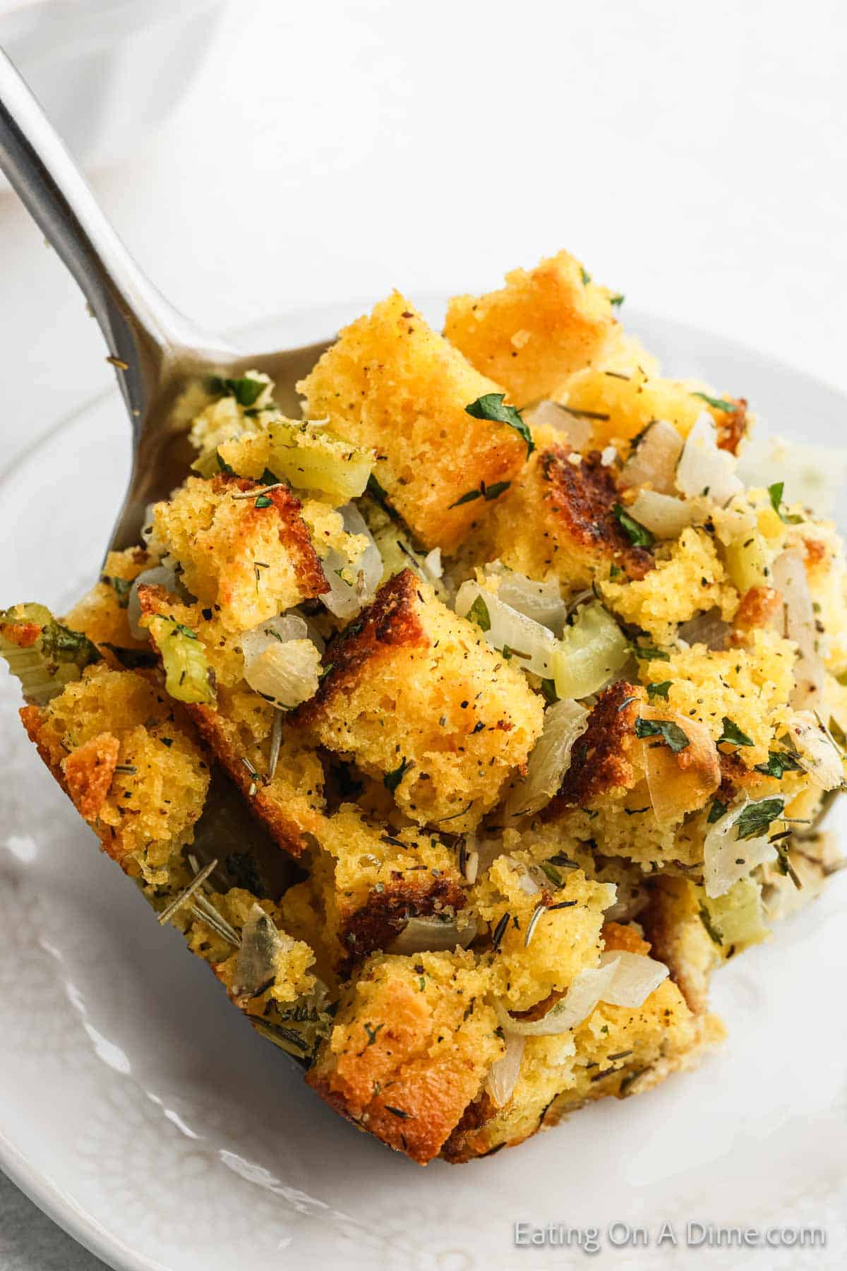 A spoonful of delicious cornbread stuffing with chopped onions, celery, and herbs is temptingly placed on a white plate. The golden-brown stuffing is moist, with visible seasoning and toasted edges that promise a burst of flavor.