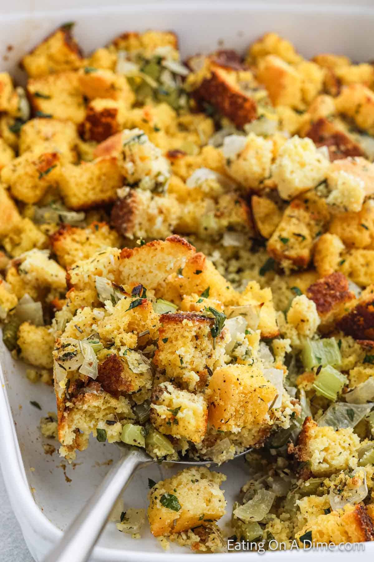 A close-up of golden brown cornbread stuffing in a baking dish features inviting chunks of cornbread, herbs, and visible pieces of onion and celery. A serving spoon is gracefully scooping some from the dish.