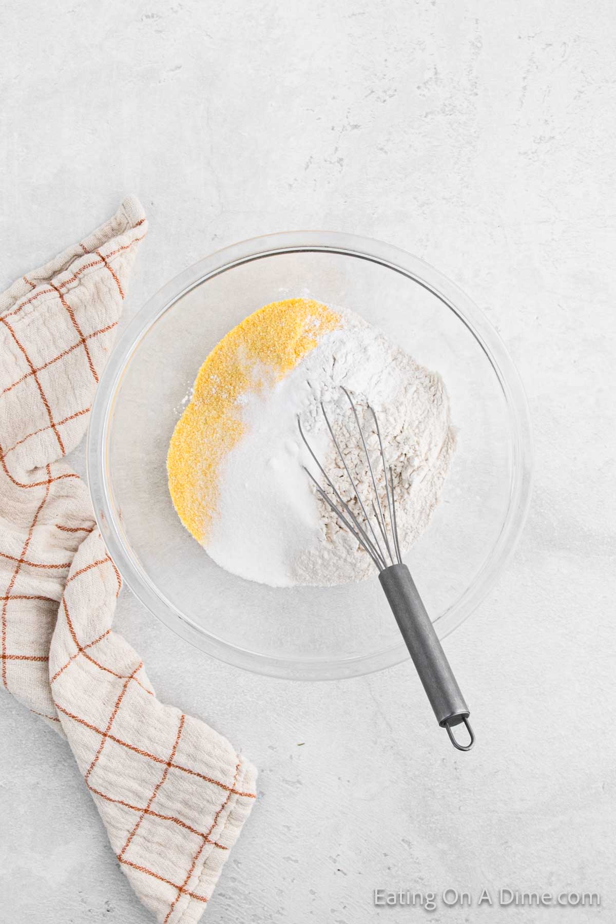 A glass bowl with flour, cornmeal, and baking powder sits on a light gray surface, ready to transform into delicious cornbread. A metal whisk rests inside the bowl, while a cream and red checkered cloth is placed beside it.