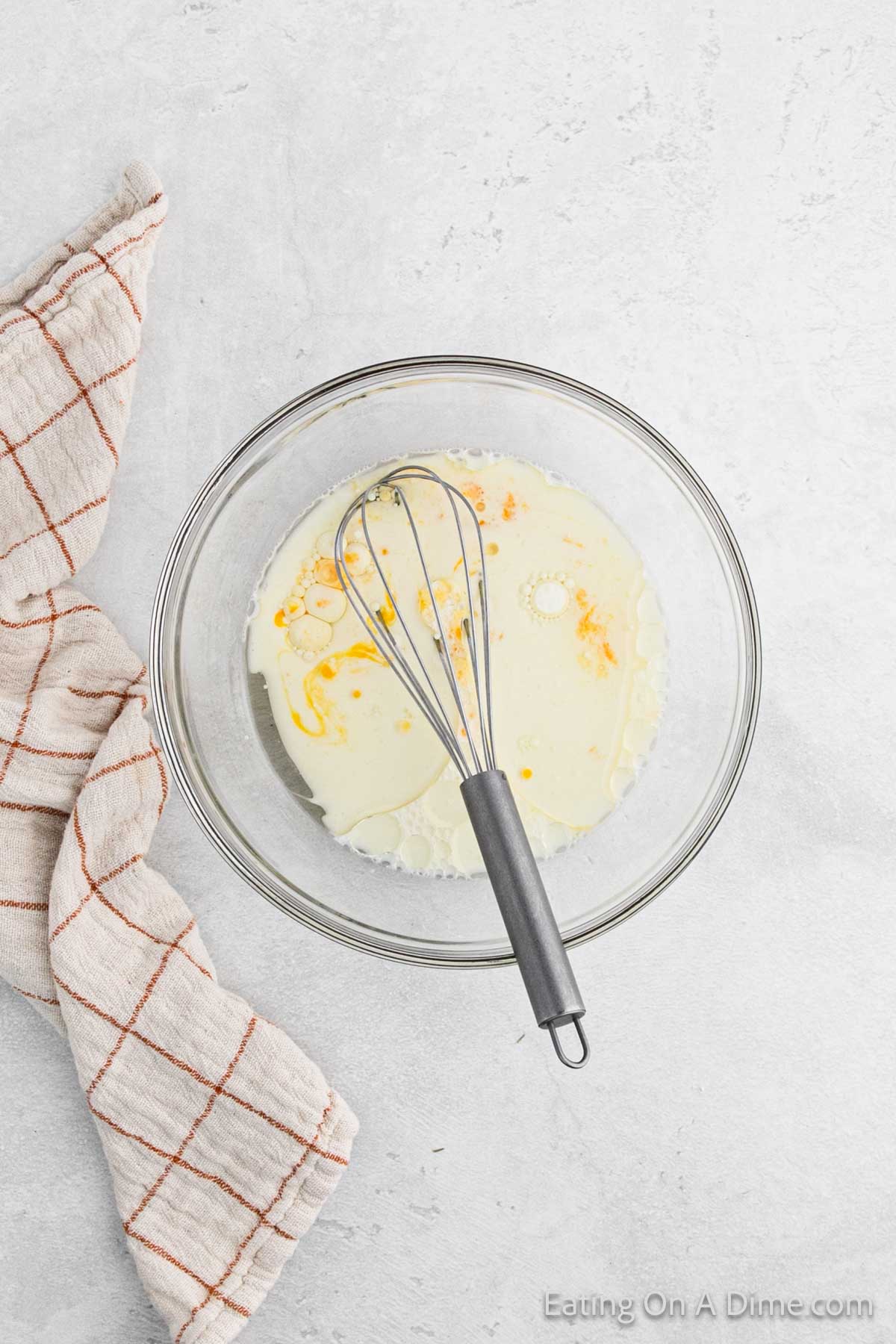 A clear glass bowl with cream and an egg yolk sits on a light gray surface, ready for mixing into cornbread. A metal whisk rests inside the bowl. Beside it, a beige kitchen towel with a red grid pattern is draped elegantly.