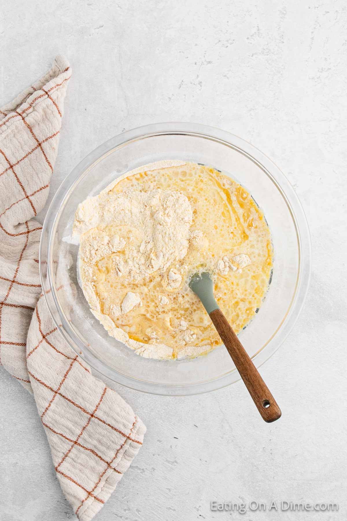 A glass bowl with ingredients for cornbread batter, including flour, eggs, and liquid, being mixed with a spatula. A beige and red checkered cloth is next to the bowl on a light gray surface.