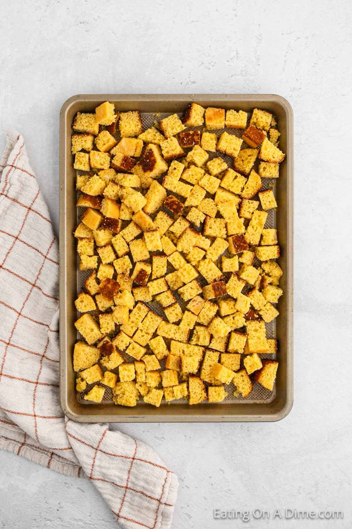 A baking sheet filled with evenly spread, diced cornbread pieces, perfect for stuffing. A white and red checkered kitchen towel is placed beside the sheet on a light gray surface.