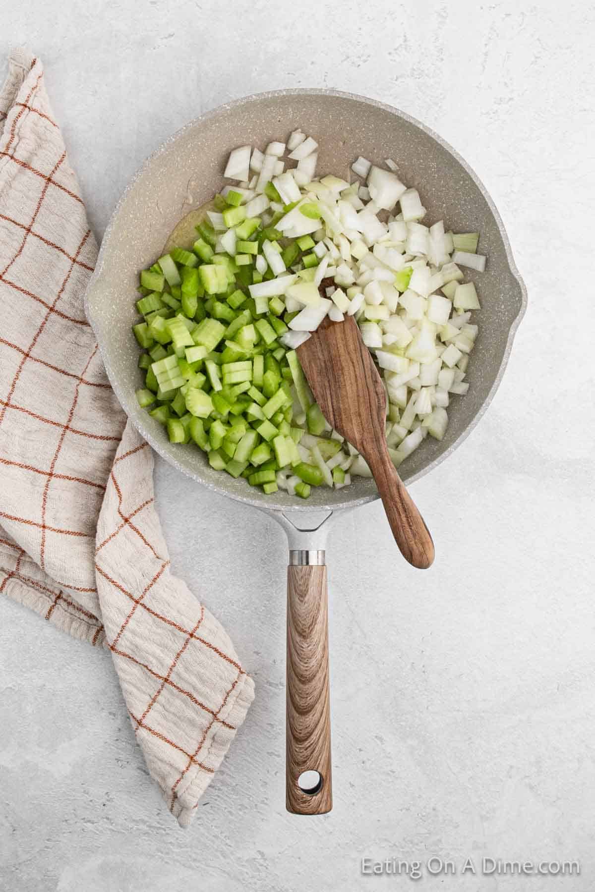 A frying pan with chopped onions and celery for the cornbread stuffing, accompanied by a wooden spatula. A beige cloth with a red grid pattern is beside the pan on a light gray surface.