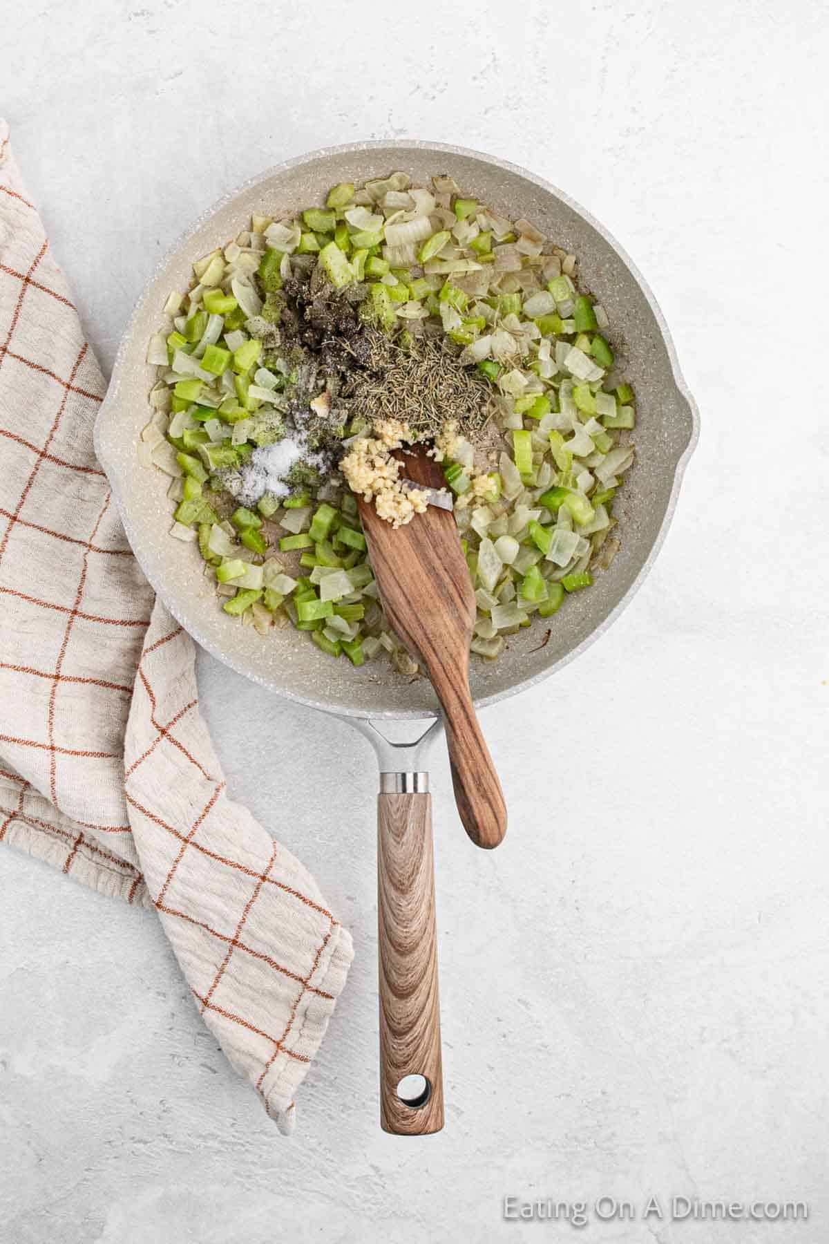 A skillet containing chopped celery, onions, minced garlic, and herbs sizzles invitingly, a wooden spatula resting nearby. Perfect for stuffing or pairing with fresh cornbread, the vibrant scene is set on a light surface next to a checked cloth.