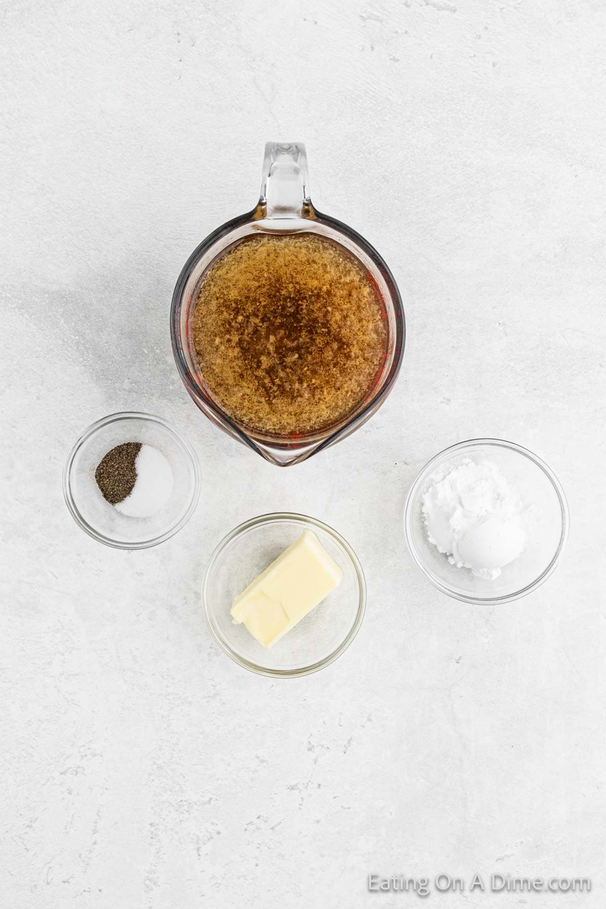 A measuring cup filled with brown sugar is surrounded by small bowls containing salt and pepper, a stick of butter, and a white powder on a light textured surface, perfect for crafting the ultimate gravy for your turkey dinner.