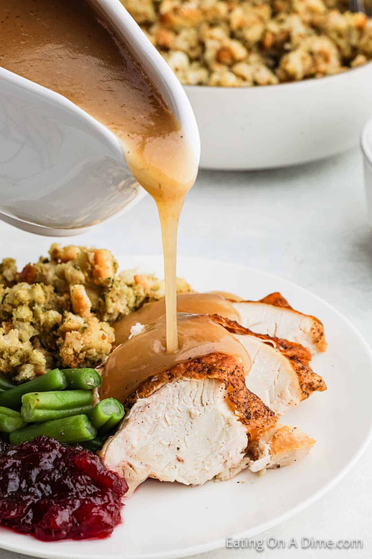 A white plate with sliced roasted turkey topped with rich, gluten-free turkey gravy being poured. The plate also includes green beans, stuffing, and cranberry sauce. A bowl of extra stuffing is visible in the background.