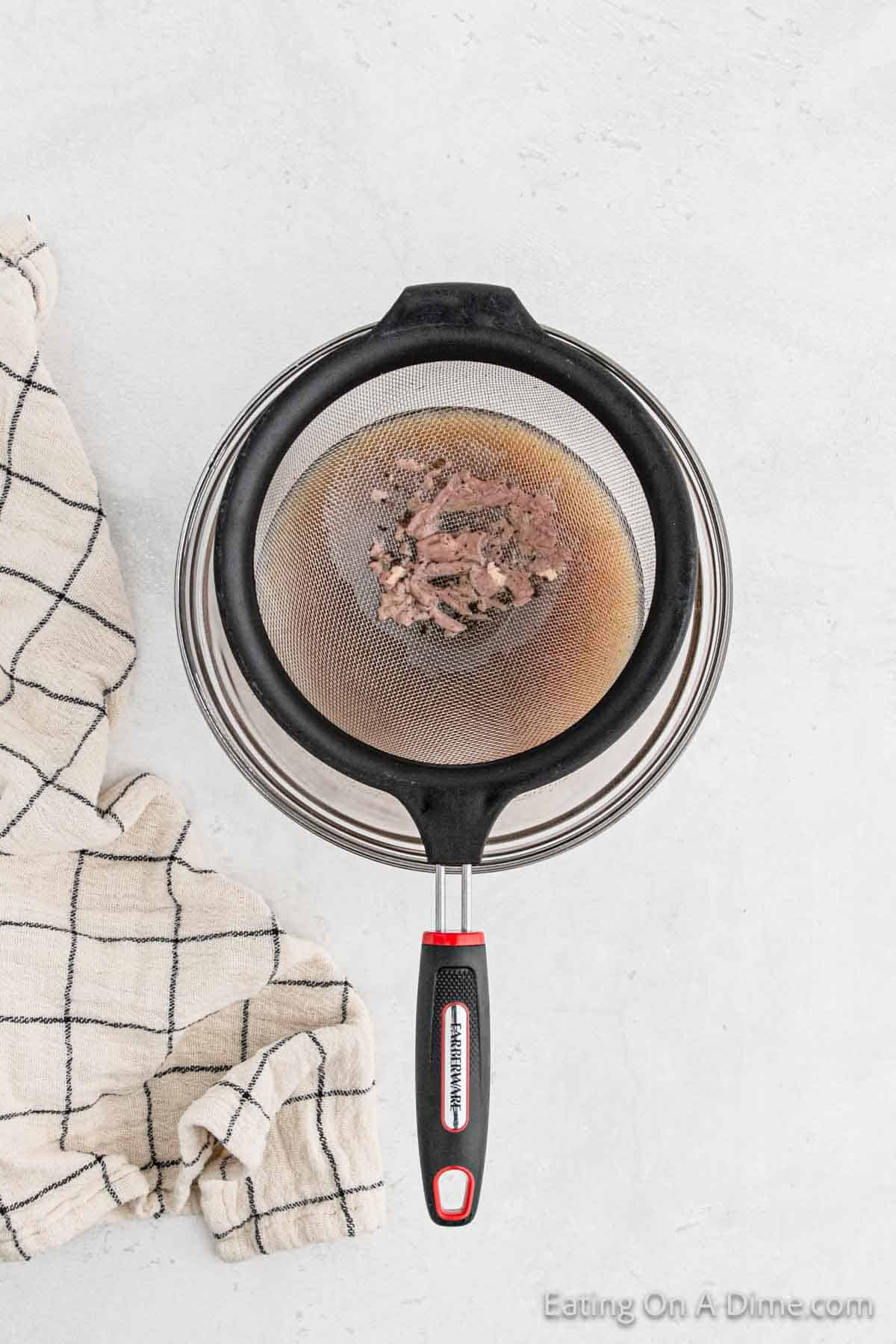 A black strainer with cooked ground beef is perched over a stainless steel bowl on a light countertop, while a white and black checkered kitchen towel rests nearby. Perfect for preparing gluten-free dishes or as a base for hearty turkey gravy.