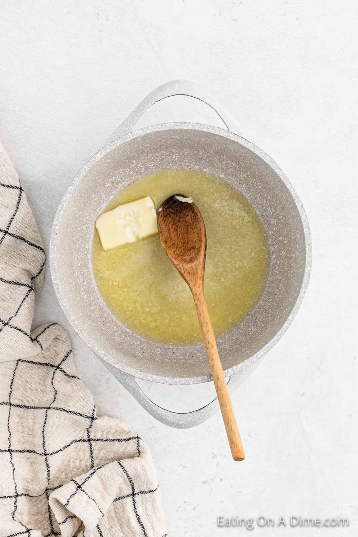 A pot of melted butter with a wooden spoon inside, placed on a light countertop, simmers next to gluten-free gravy. To the side, there's a cloth with a checkered pattern.