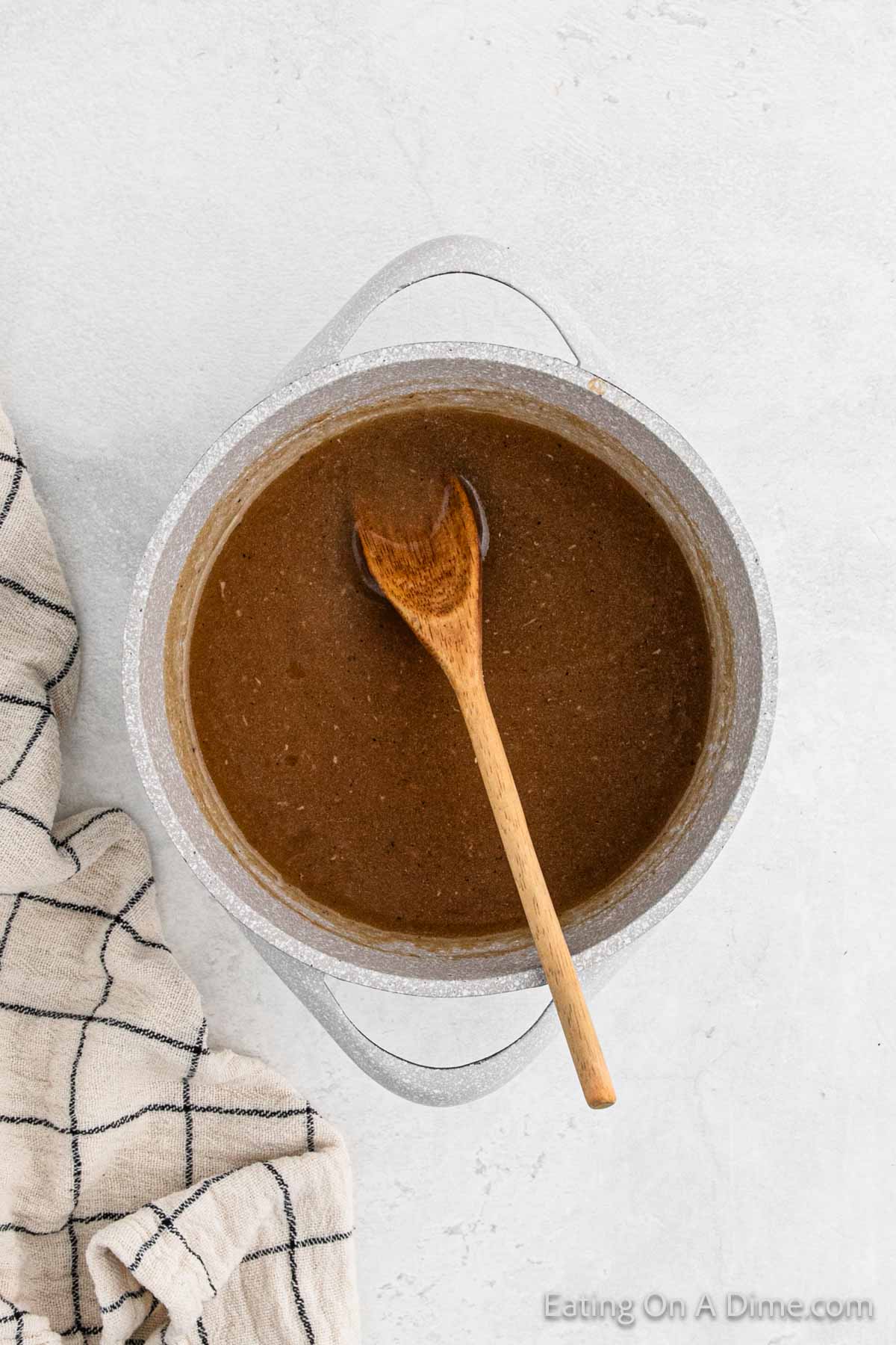 A pot of rich brown gravy, perfect for turkey, sits on a light surface with a wooden spoon inside. A checked cloth is laid beside the pot, ready for a delightful meal.