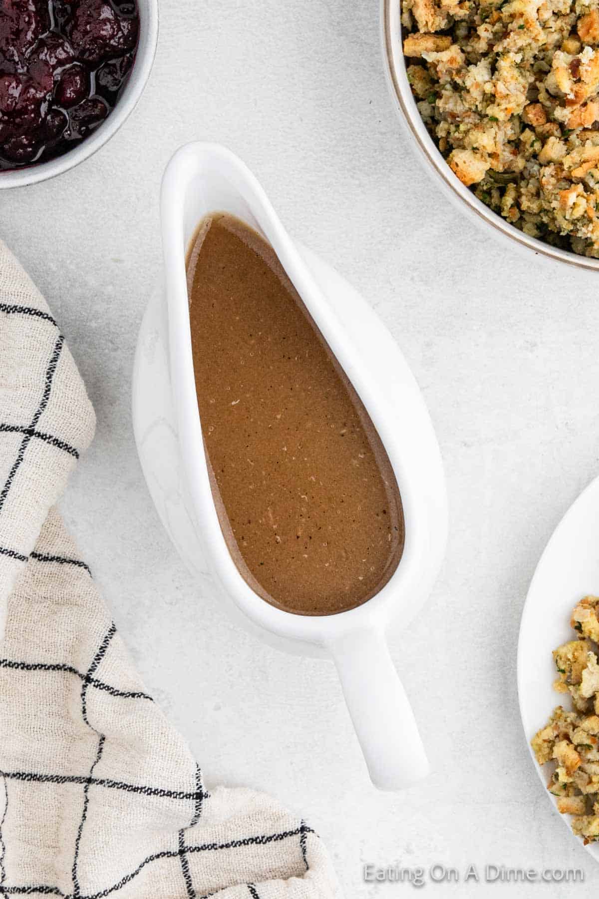 A white gravy boat brimming with rich, gluten-free brown gravy is surrounded by a beige dish towel, alongside a bowl of stuffing and a dish of cranberry sauce on a light gray surface. Perfect for your turkey feast.