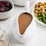 A white gravy boat filled with rich, brown gluten-free gravy sits on a table, surrounded by a bowl of cranberry sauce and a plate of mixed vegetables and stuffing. The light background complements the scene, while a folded napkin is partially visible, hinting at the awaiting turkey feast.