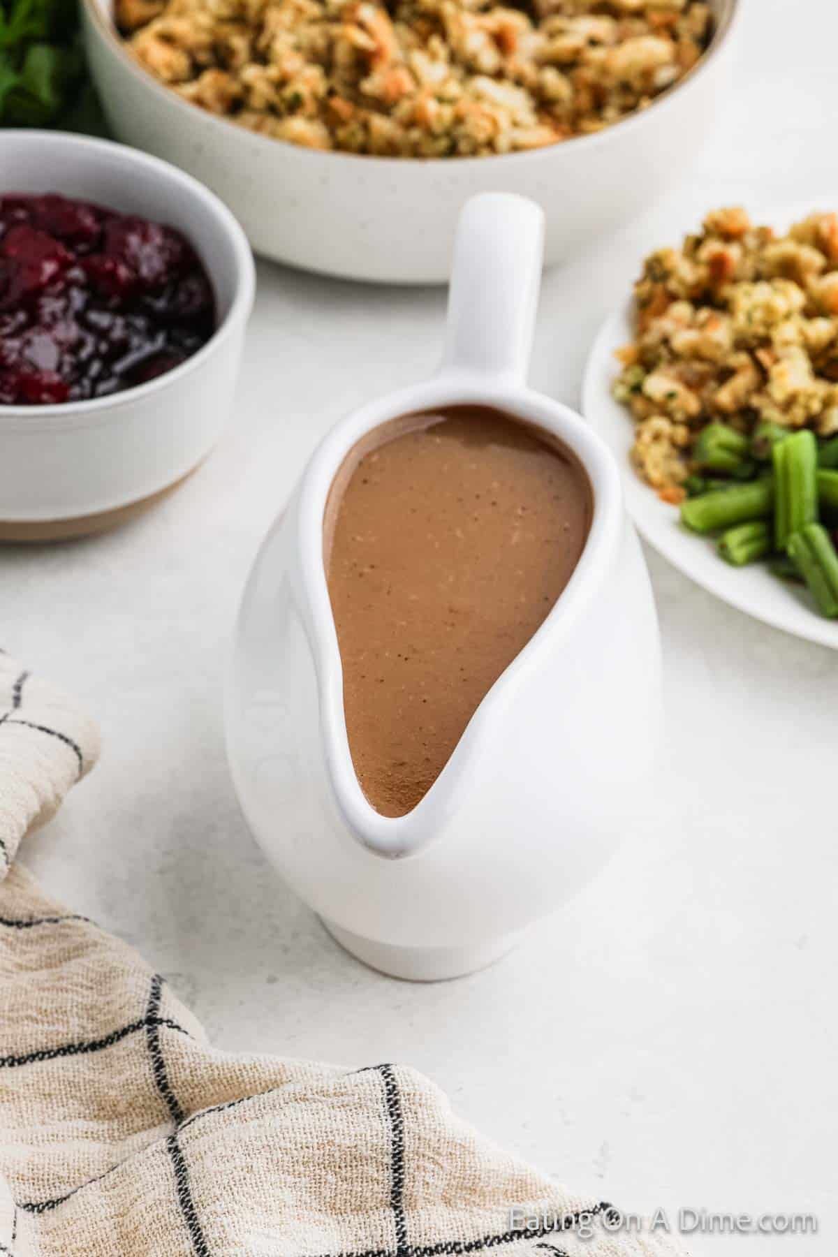 A white gravy boat brimming with rich, gluten-free turkey gravy sits on a table. Encircling it are dishes of cranberry sauce, stuffing, and green beans atop a checkered cloth. The light, neutral background enhances the inviting setting. 