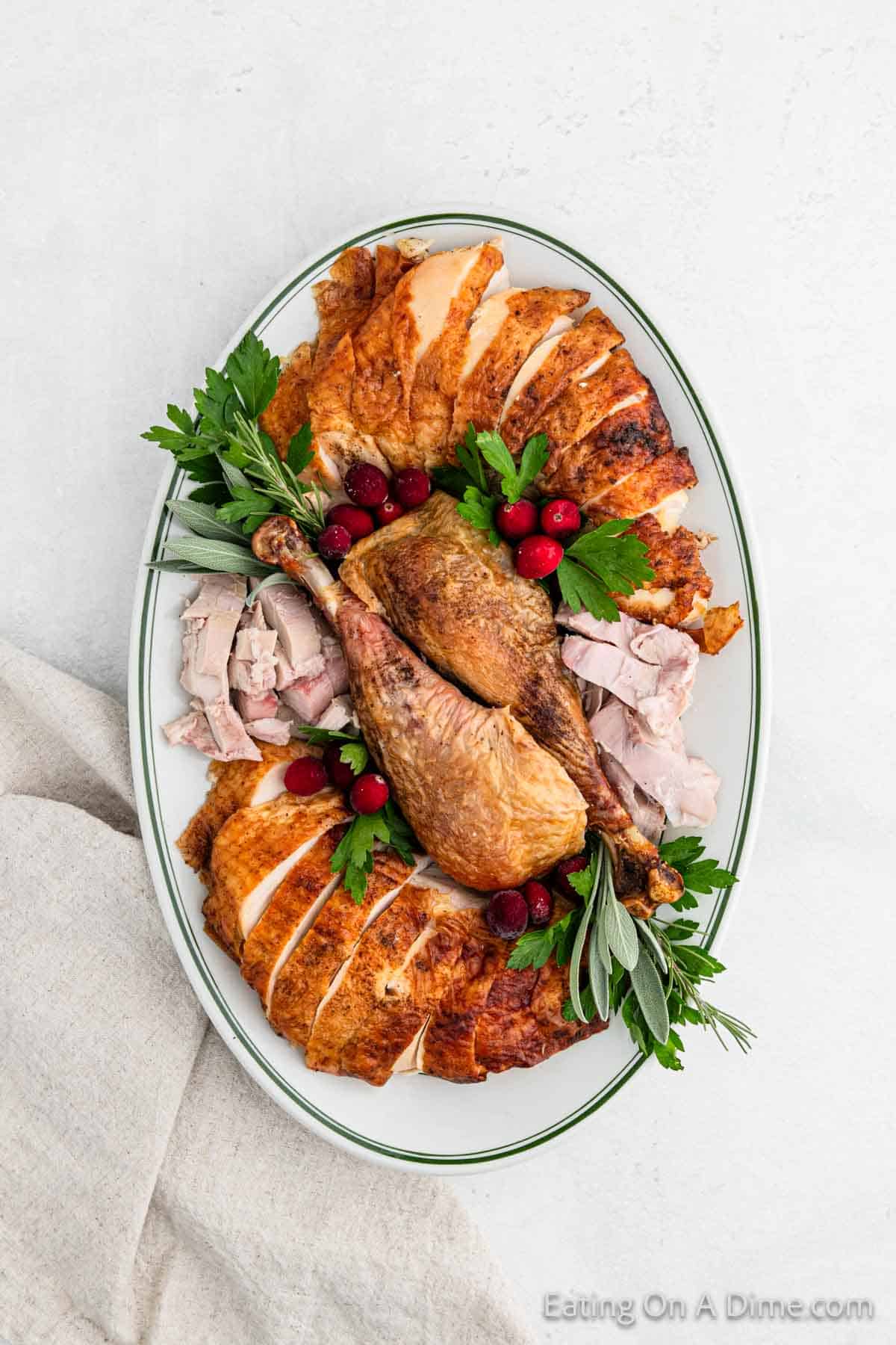 A platter of expertly carved turkey garnished with fresh parsley, sage, and cranberries. The arranged slices encircle two drumsticks at the center. This beautifully presented dish rests on a white oval plate with a green rim, set on a light surface, showcasing perfect turkey carving skills.