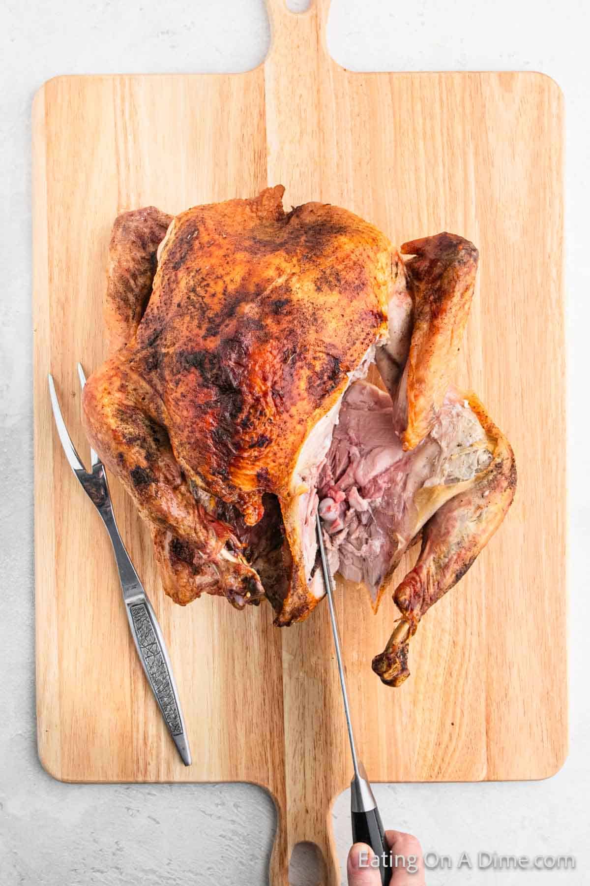 A roasted turkey with crispy skin is being expertly carved on a wooden cutting board. Showcasing refined carving techniques, a meat fork rests beside the turkey as the knife reveals juicy slices, perfecting the art of turkey preparation.