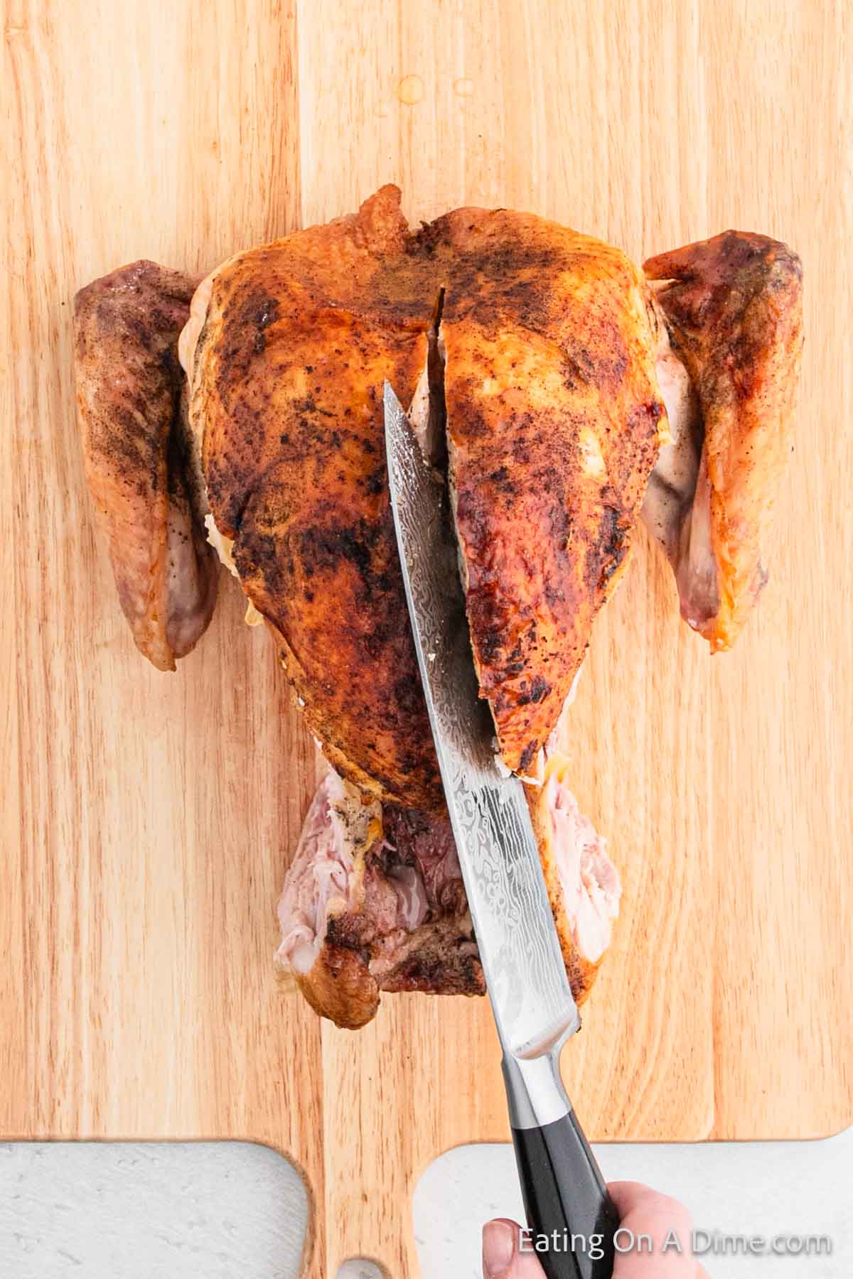 A whole roasted chicken is placed on a wooden cutting board. A hand holds a knife, demonstrating how to carve along the breastbone. The golden-brown skin is perfectly seasoned, and a corner of the cutting board is visible at the bottom, reminiscent of turkey carving techniques.