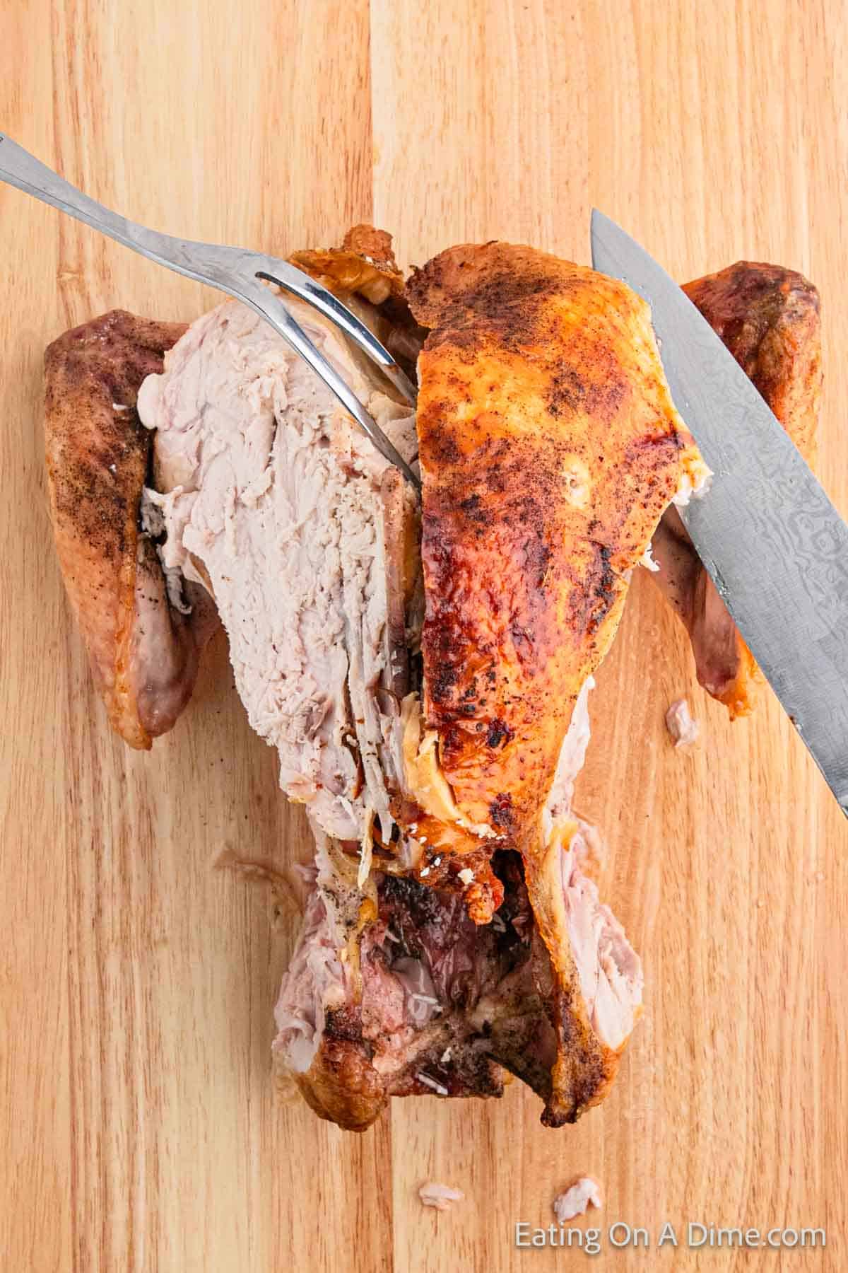 A knife and fork expertly carve a roasted chicken on a wooden cutting board. The crispy, brown skin gives way to tender white meat beneath. In the background, a subtle "Eating On A Dime" watermark complements this turkey carving demonstration.