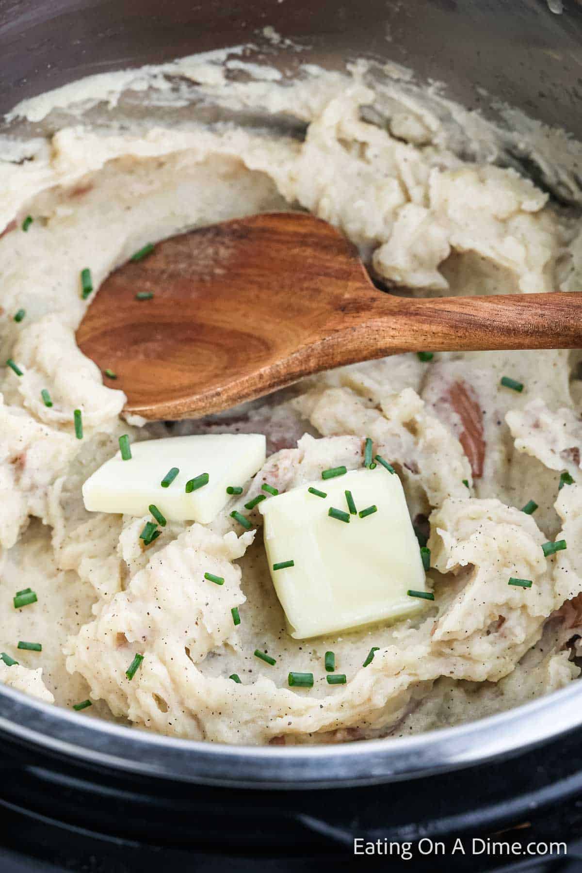 A pot of creamy mashed red potatoes with specks of pepper and chopped chives, topped with two pats of melting butter. A wooden spoon rests on the side, ready to serve this comforting dish straight from the Instant Pot.