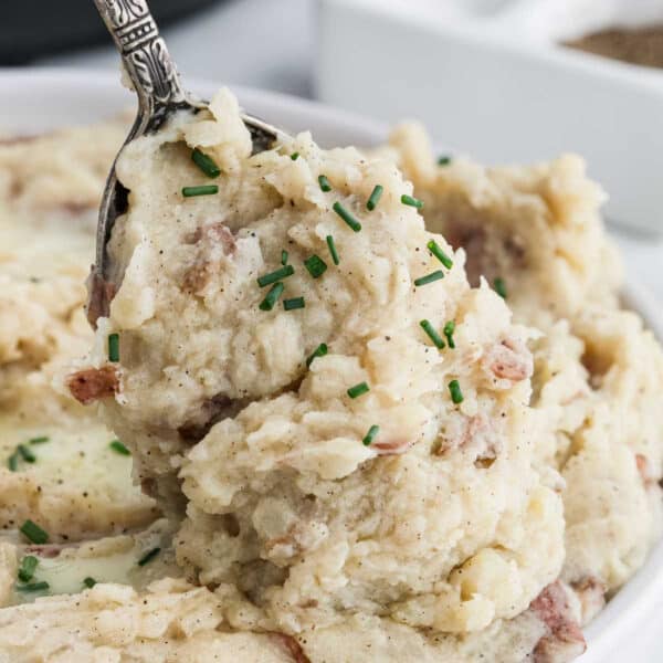 Close-up of a spoon lifting creamy mashed red potatoes from a bowl, garnished with chopped chives and black pepper. Using an Instant Pot makes this delicious side dish effortless and perfect every time.