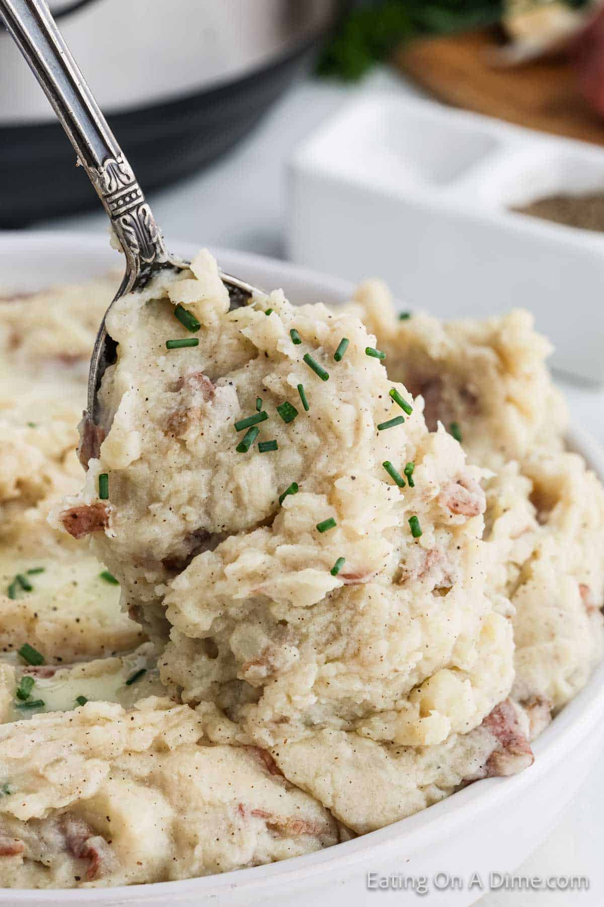 A close-up of creamy homemade mashed red potatoes being scooped with a silver spoon. Garnished with chopped chives, the mash has visible pieces of red-skinned potatoes, adding a rustic texture to this classic dish.