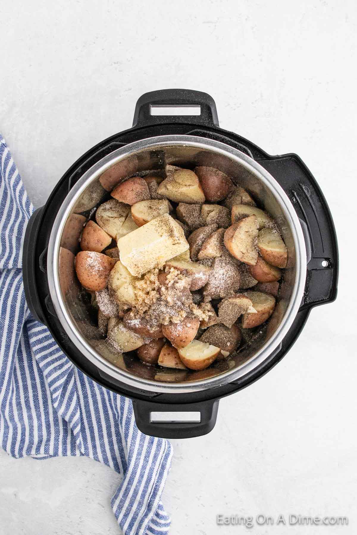 An Instant Pot filled with cubed red potatoes topped with butter, garlic, and seasonings. A blue and white striped cloth is placed beside the pot on a light gray surface.