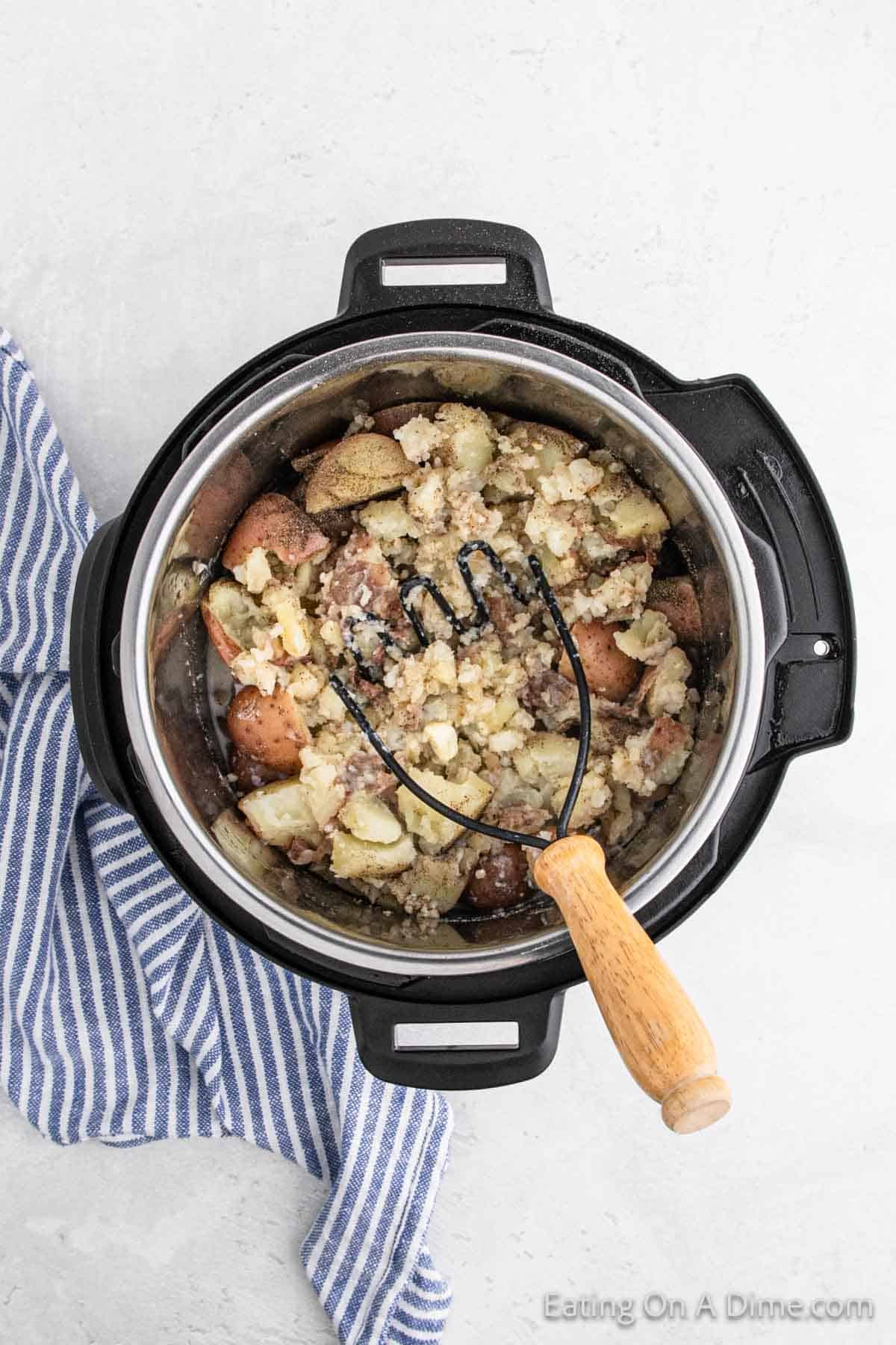 A pot of creamy mashed potatoes with a potato masher inside sits on a light surface. Beside it, a blue and white striped cloth adds a touch of charm. The comforting sight invites you to enjoy the warmth and flavor of freshly mashed red potatoes.