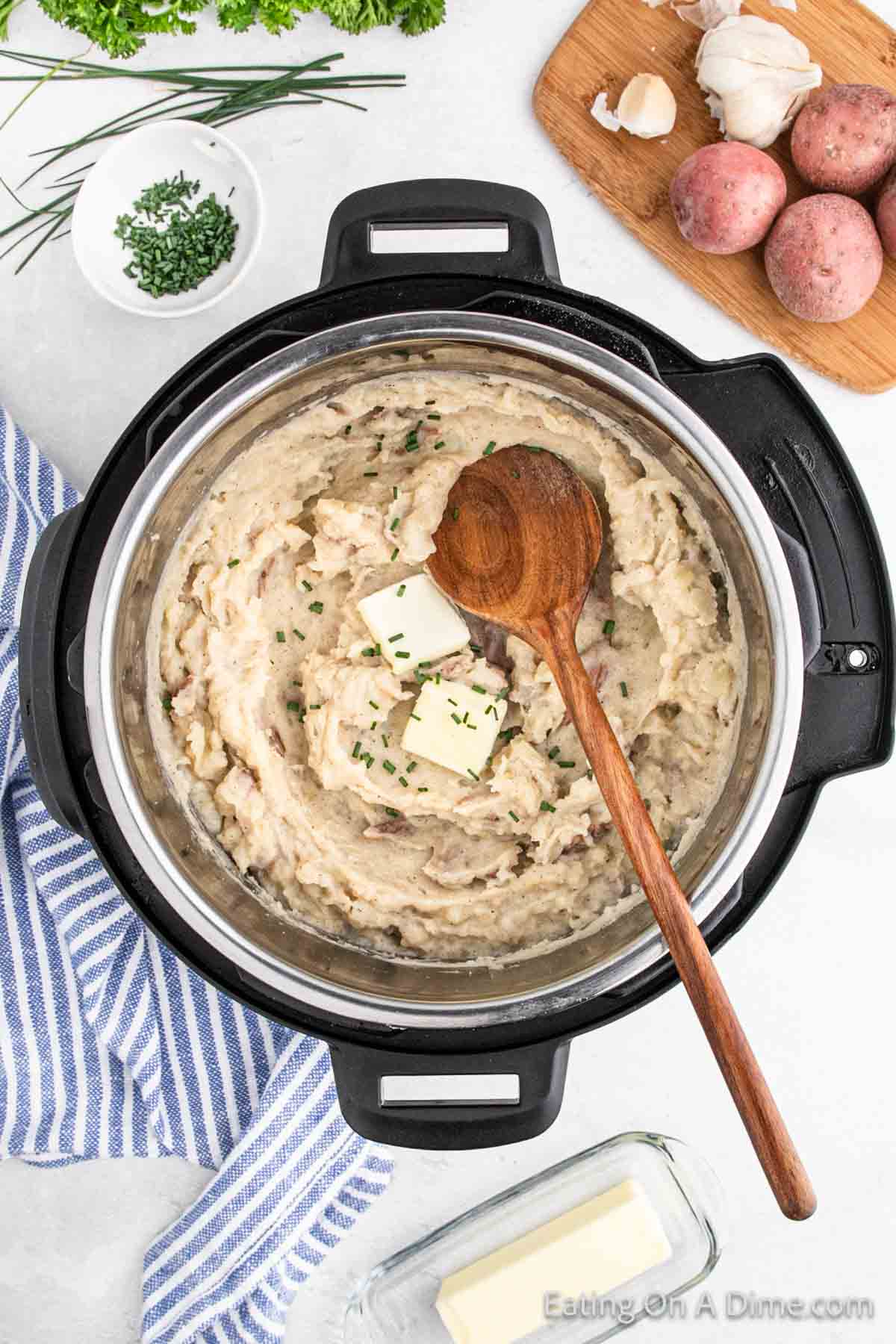 A pot of creamy mashed red potatoes garnished with chives and two pats of butter, with a wooden spoon resting on top. Surrounding items include a blue striped kitchen towel, a cutting board with garlic and more red potatoes, and a small bowl with herbs.