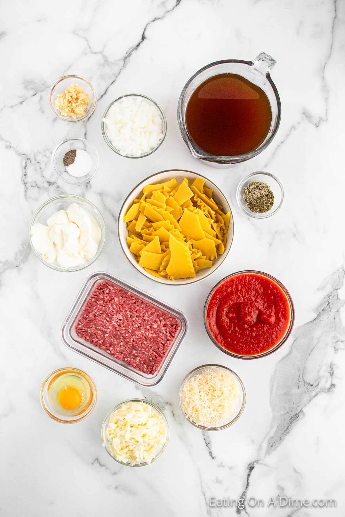 Top view of ingredients on a marble surface for a delicious lasagna soup: chopped garlic, minced onion, beef broth, Italian seasoning, salt and pepper, sour cream, broken lasagna noodles, ground beef, crushed tomatoes, an egg, shredded mozzarella, and Parmesan cheese.