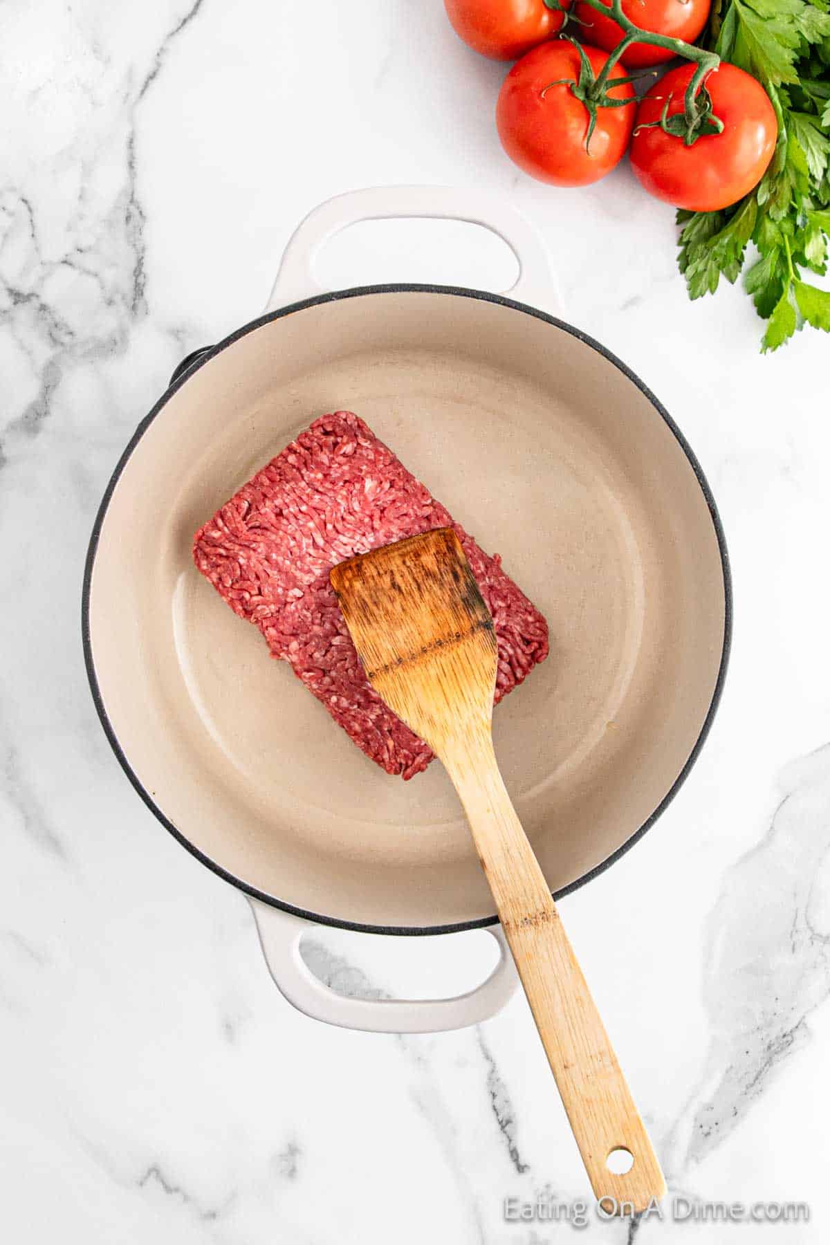 A delicious lasagna base is created as raw ground beef sizzles in a beige pot on a marble countertop, stirred gently with a wooden spatula. Fresh tomatoes and parsley wait nearby, ready to enhance this savory dish.