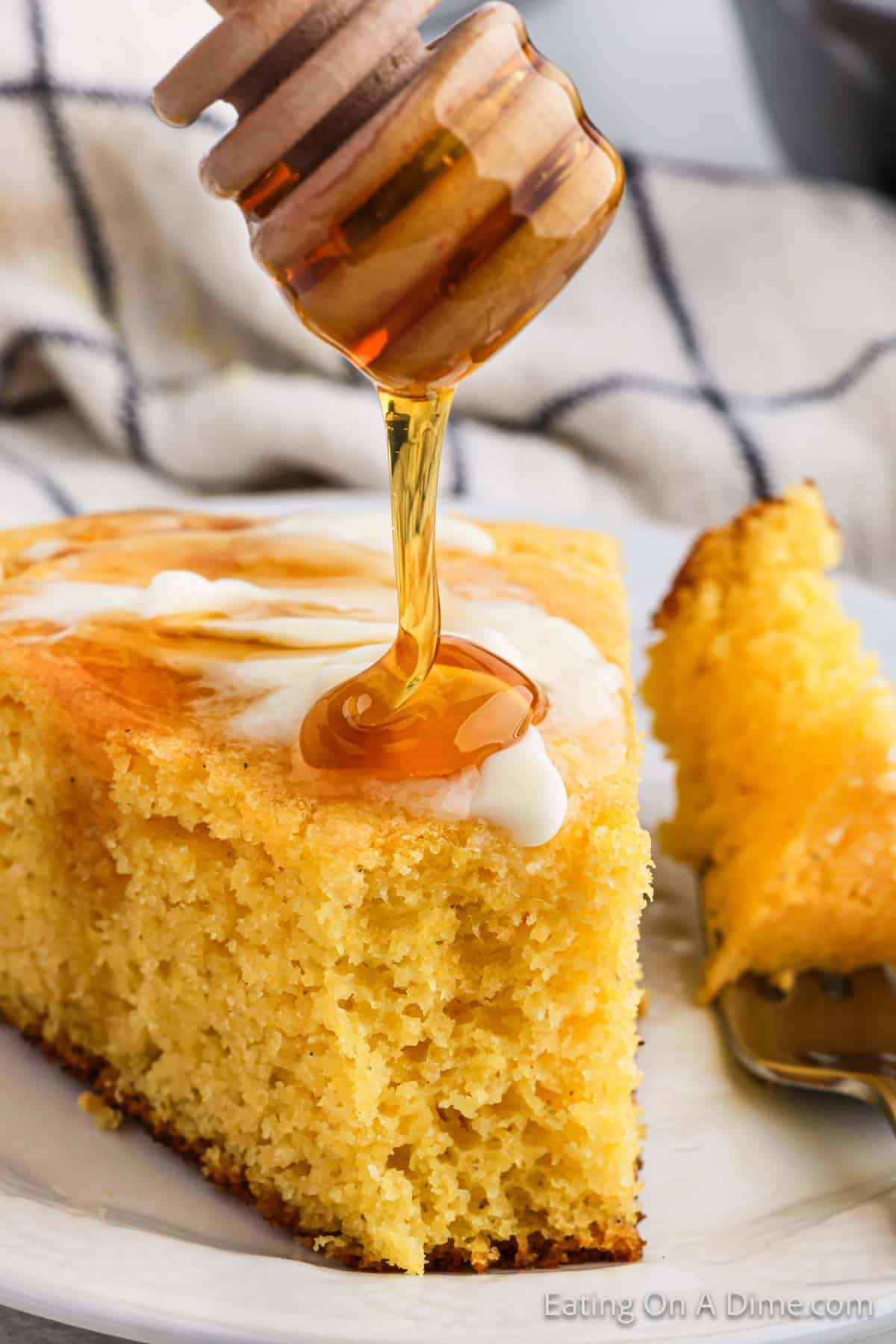 A slice of moist southern cornbread is topped with a drizzle of honey being poured from a wooden honey dipper. A fork rests beside the cornbread recipe on a white plate, and a striped cloth is visible in the background.