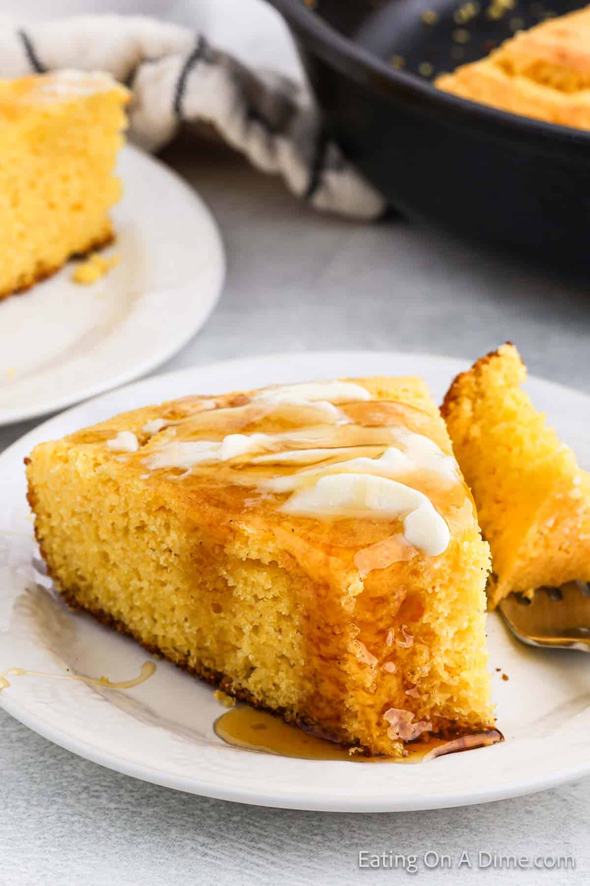 A slice of Southern cornbread with a drizzle of honey and a pat of butter melting on top. It is served on a white plate with a fork, and another piece of this classic Southern recipe is visible in the background on a separate plate.