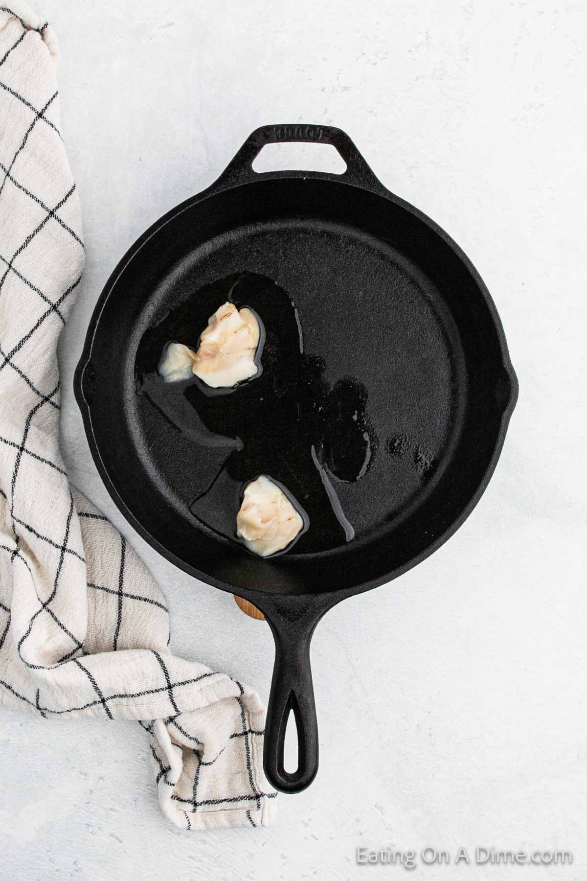 A black cast iron skillet with melting butter sits on a light surface, ready for the perfect Southern cornbread. A checked kitchen towel is partially visible beside the skillet, adding a touch of rustic charm to this Southern cooking scene.