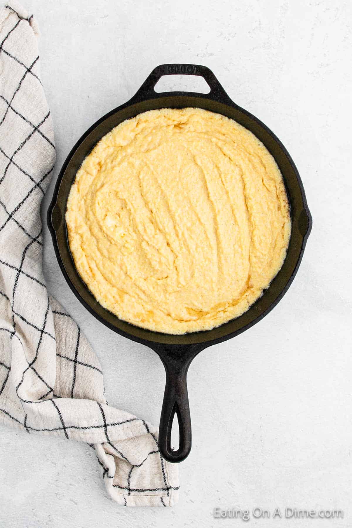 A black cast iron skillet filled with freshly baked Southern cornbread sits on a light gray surface. A white kitchen towel with a black grid pattern is draped next to it. The Southern recipe results in cornbread that appears golden and slightly textured on top.