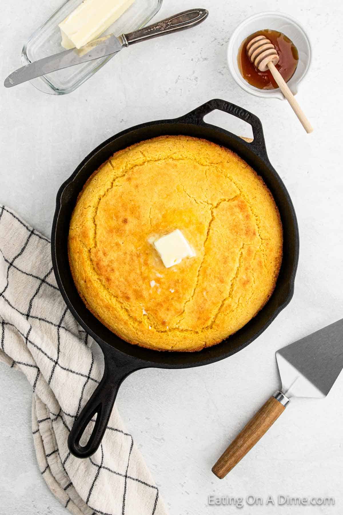 A golden Southern cornbread topped with a pat of butter sits in a black skillet. Beside it, a cutting utensil rests on a checkered cloth. In the background, there's a dish with butter and a small bowl filled with honey and a honey dipper, ready to complete this classic recipe's flavor harmony.