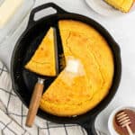 A sliced Southern cornbread in a cast-iron skillet with a pat of butter melting on top. One slice is being lifted out with a spatula. Nearby are a checkered cloth, a dish of butter, and a small bowl of honey, adding charm to this classic Southern recipe scene.